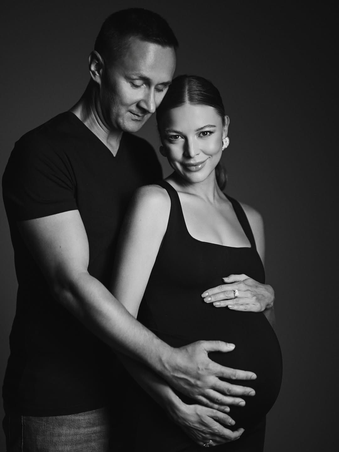 An intimate black and white studio portrait of a husband lovingly embracing his pregnant wife's belly