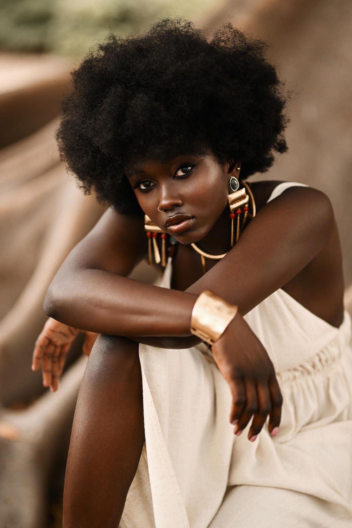 Intimate, moody close-up portrait of an elegant model resting her chin on her arms, highlighted by textured lighting and gold cuffs.