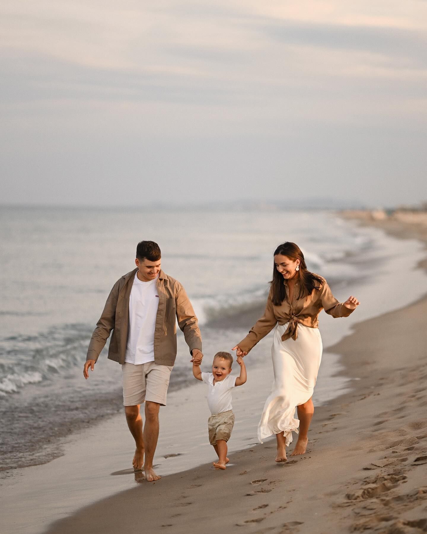 Happy parents holding hands and walking with their small child along the sandy coastline during a sunset family photoshoot in Valencia.