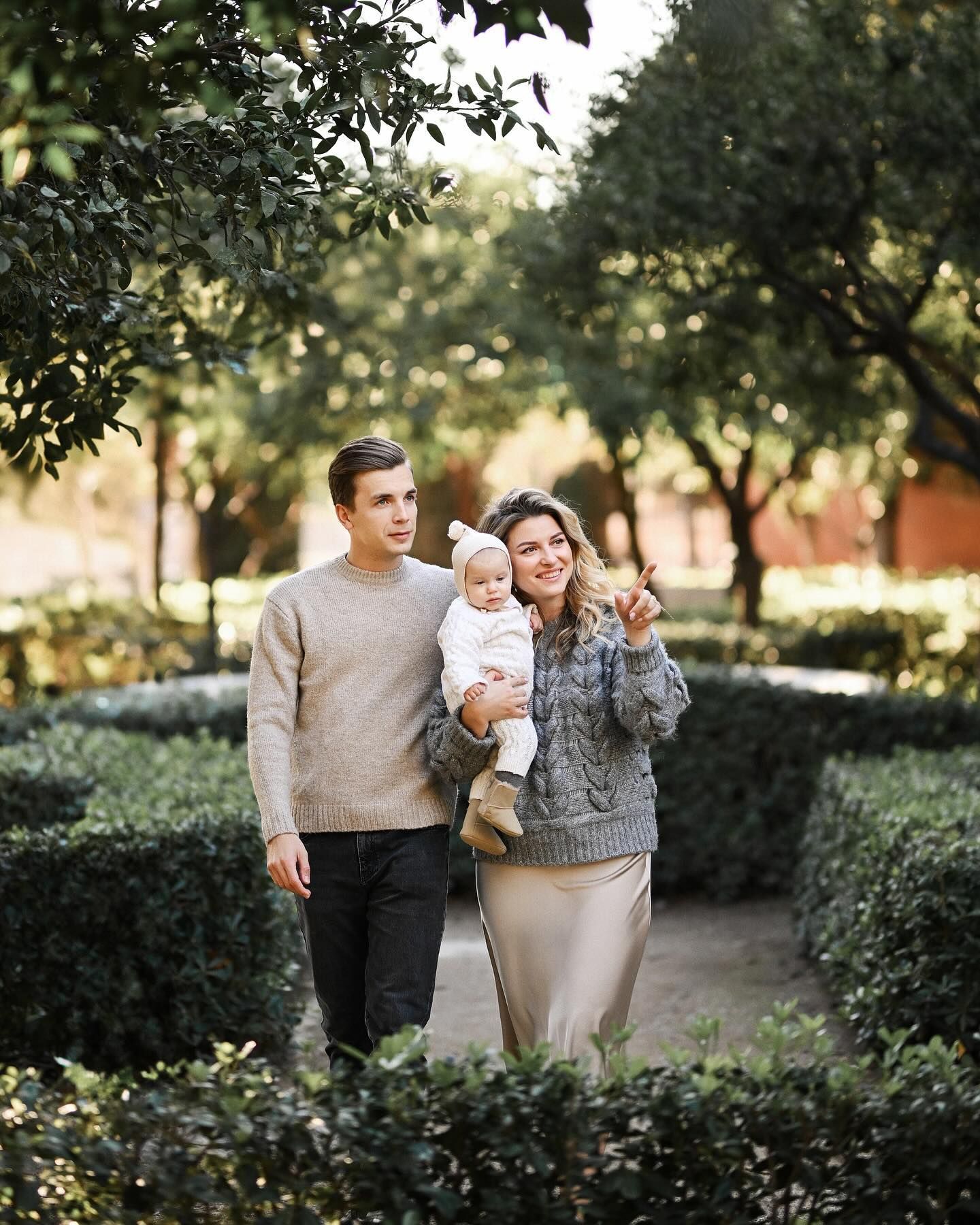 Parents holding their baby while walking along a path surrounded by green bushes in a Valencia park during a baby session.