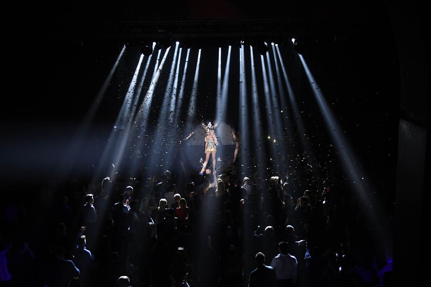 Wide shot of a live concert performance captured from above under dramatic stage lighting beams and falling glitter.