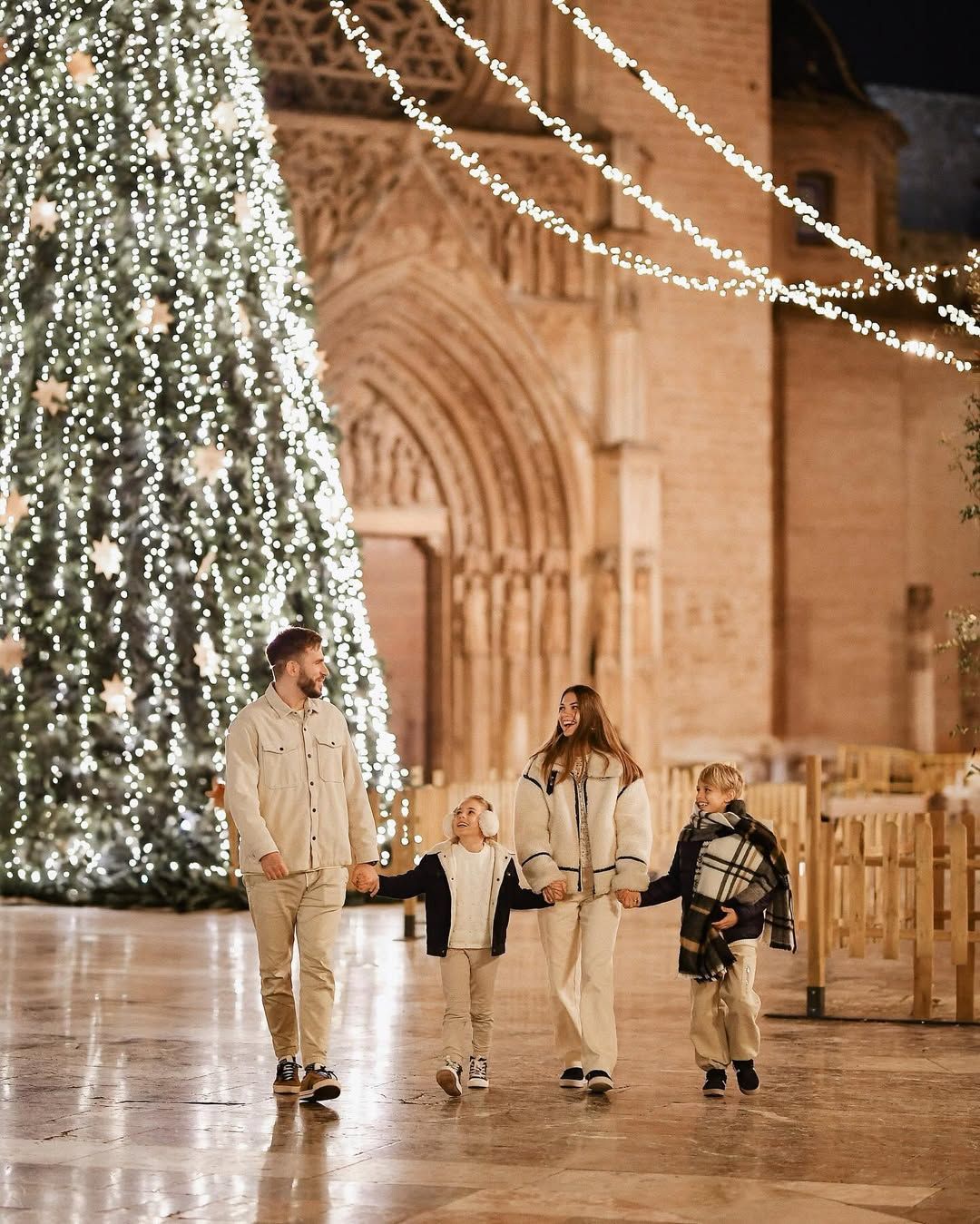 Happy family holding hands and walking joyfully near a giant illuminated Christmas tree during a holiday photoshoot in Valencia