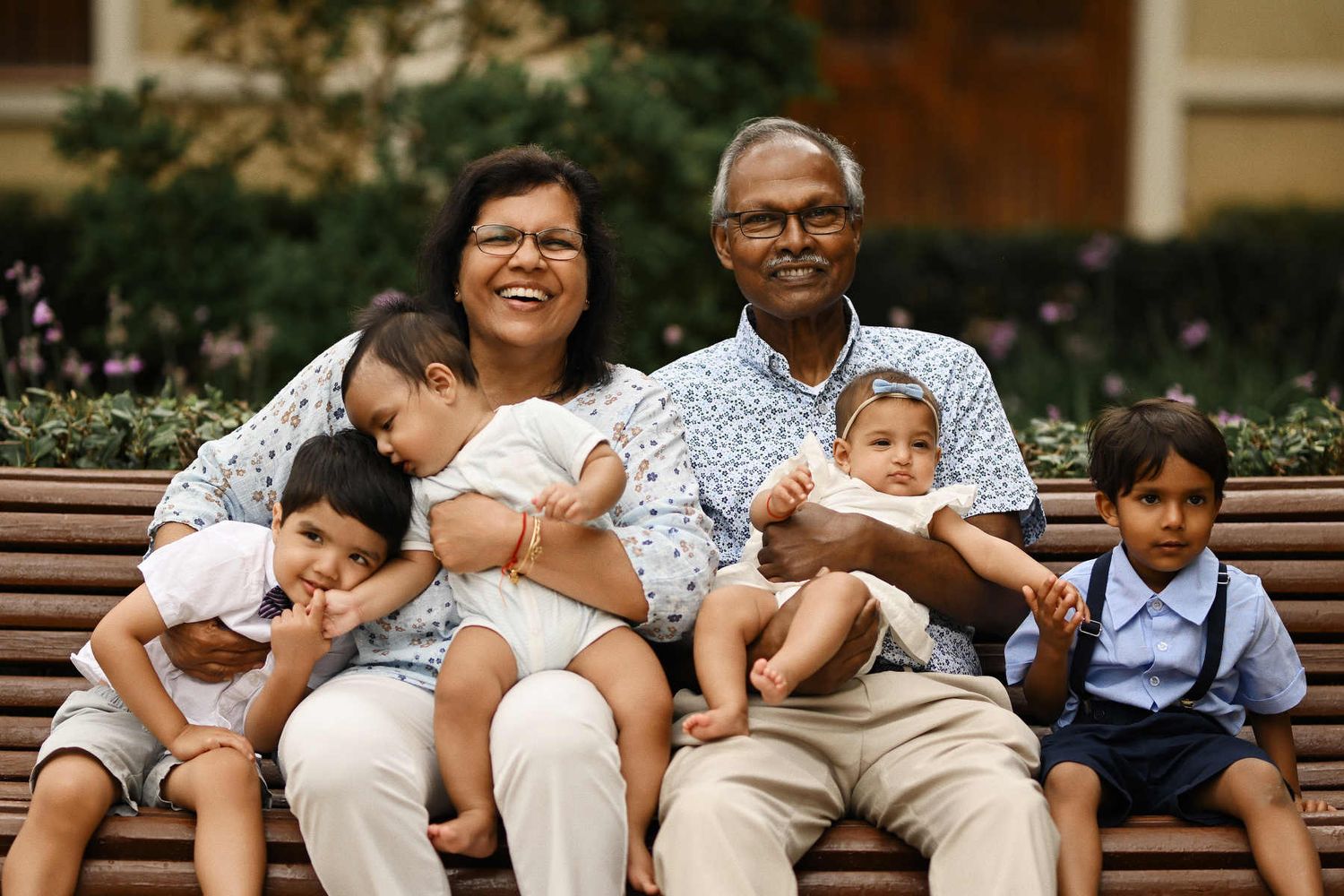 Happy grandparents sitting on a park bench, holding and smiling with their four young grandchildren during a family portrait session.