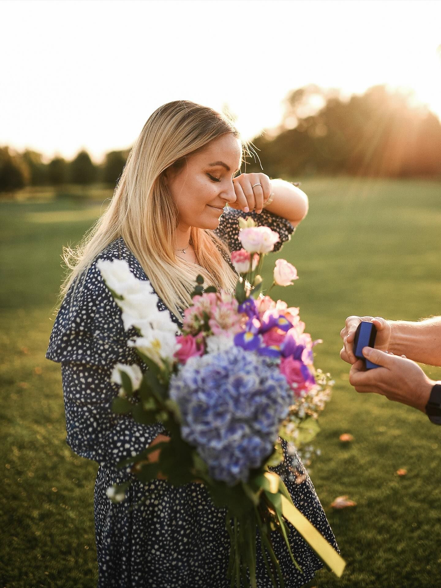Close-up of an emotional woman wiping away tears of joy while receiving a ring box from her partner in a sunlit grassy field.