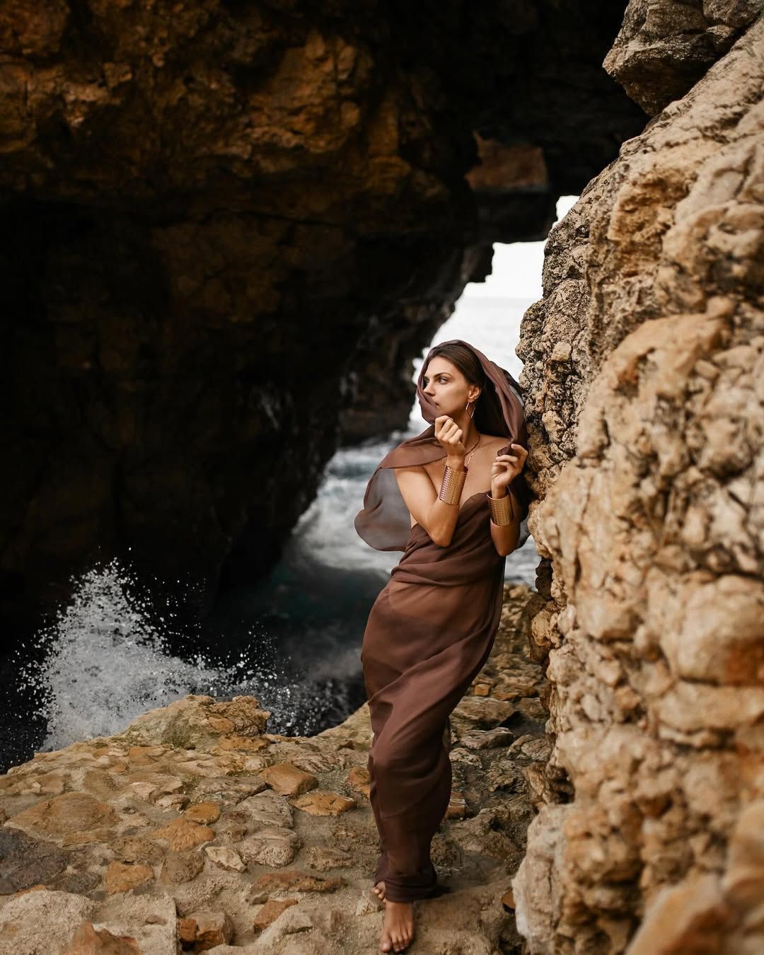A woman in an elegant brown outfit posing against dramatic cliff formations at Cala Moraig.
