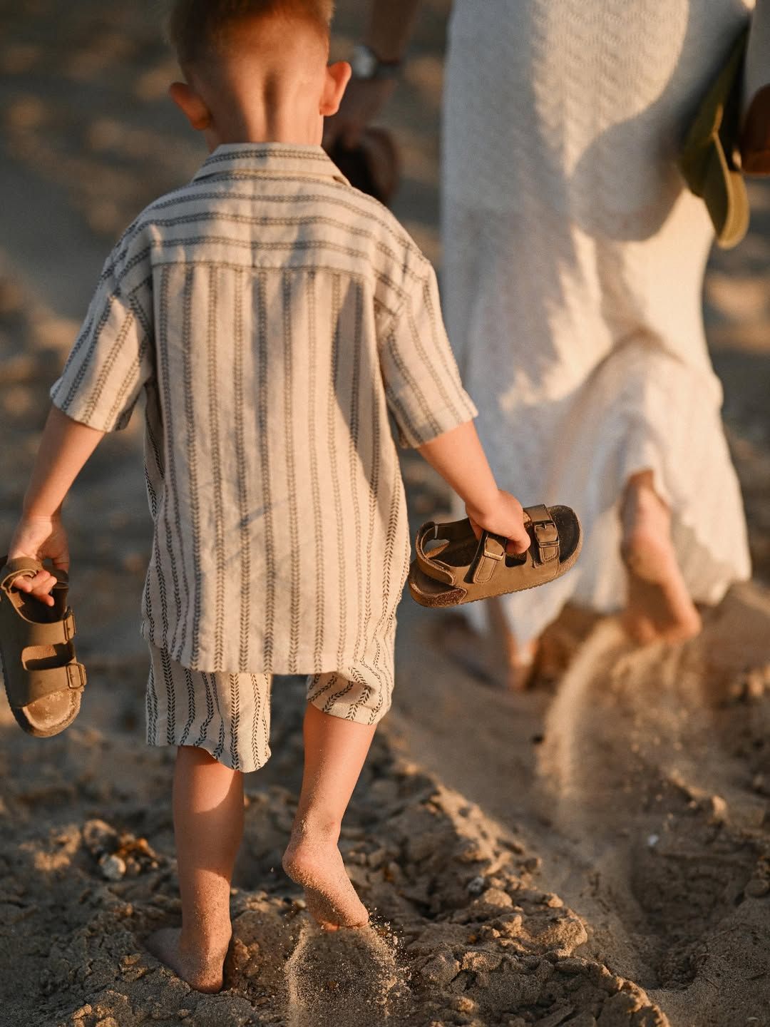 A young boy walks barefoot carrying his sandals with his mother trailing behind during a family beach session at El Saler.