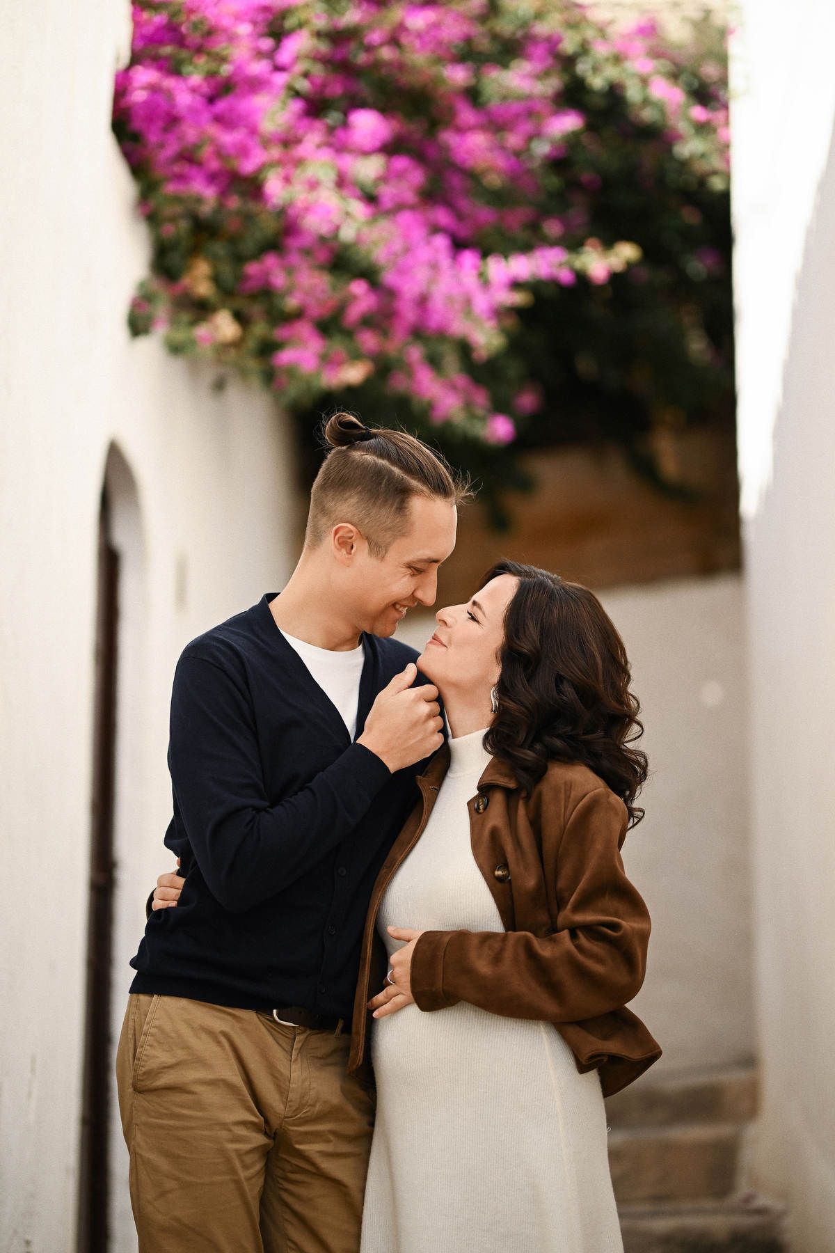 Close-up of a smiling couple expecting a baby, standing under vibrant purple flowers in the charming streets of Altea
