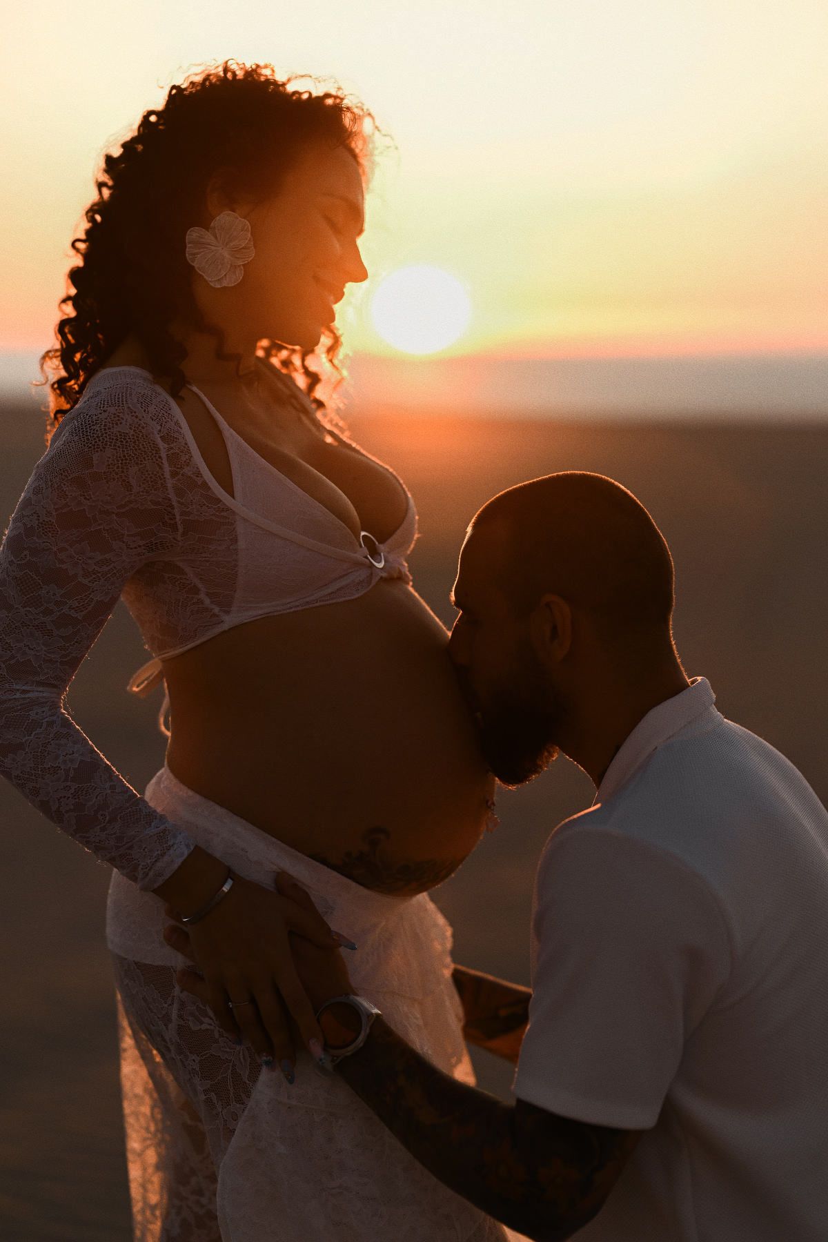 A tender moment of an expectant father kissing his pregnant partner's belly on a beach during a golden sunset.