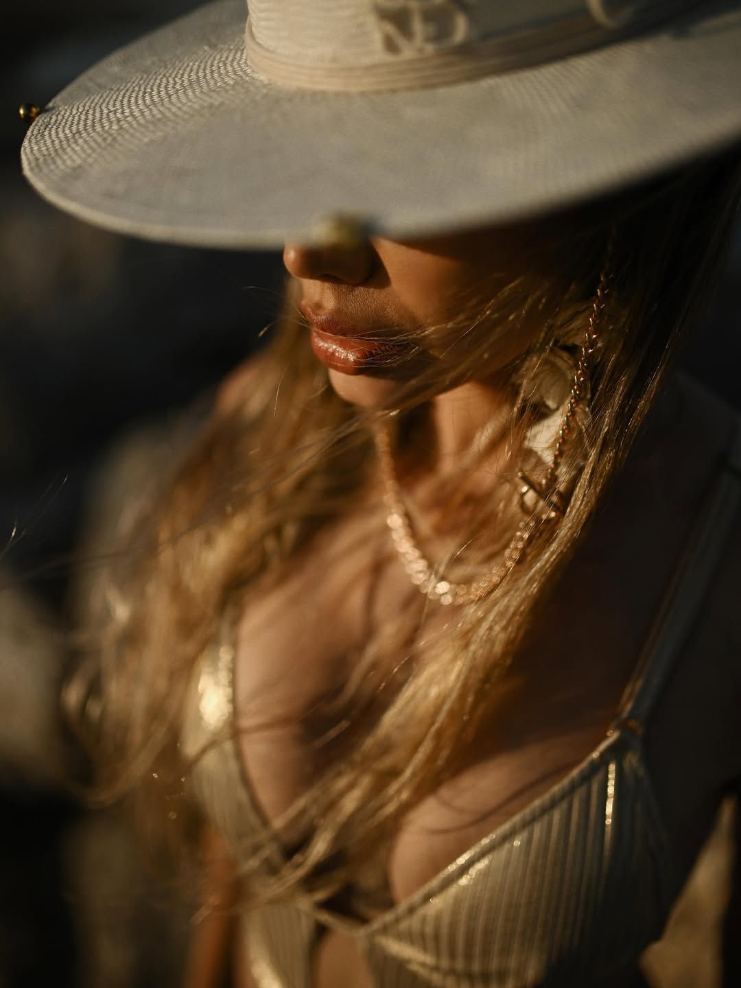 A cinematic close-up focusing on a woman's face and golden jewelry, shaded by a wide-brimmed straw hat at the beach.