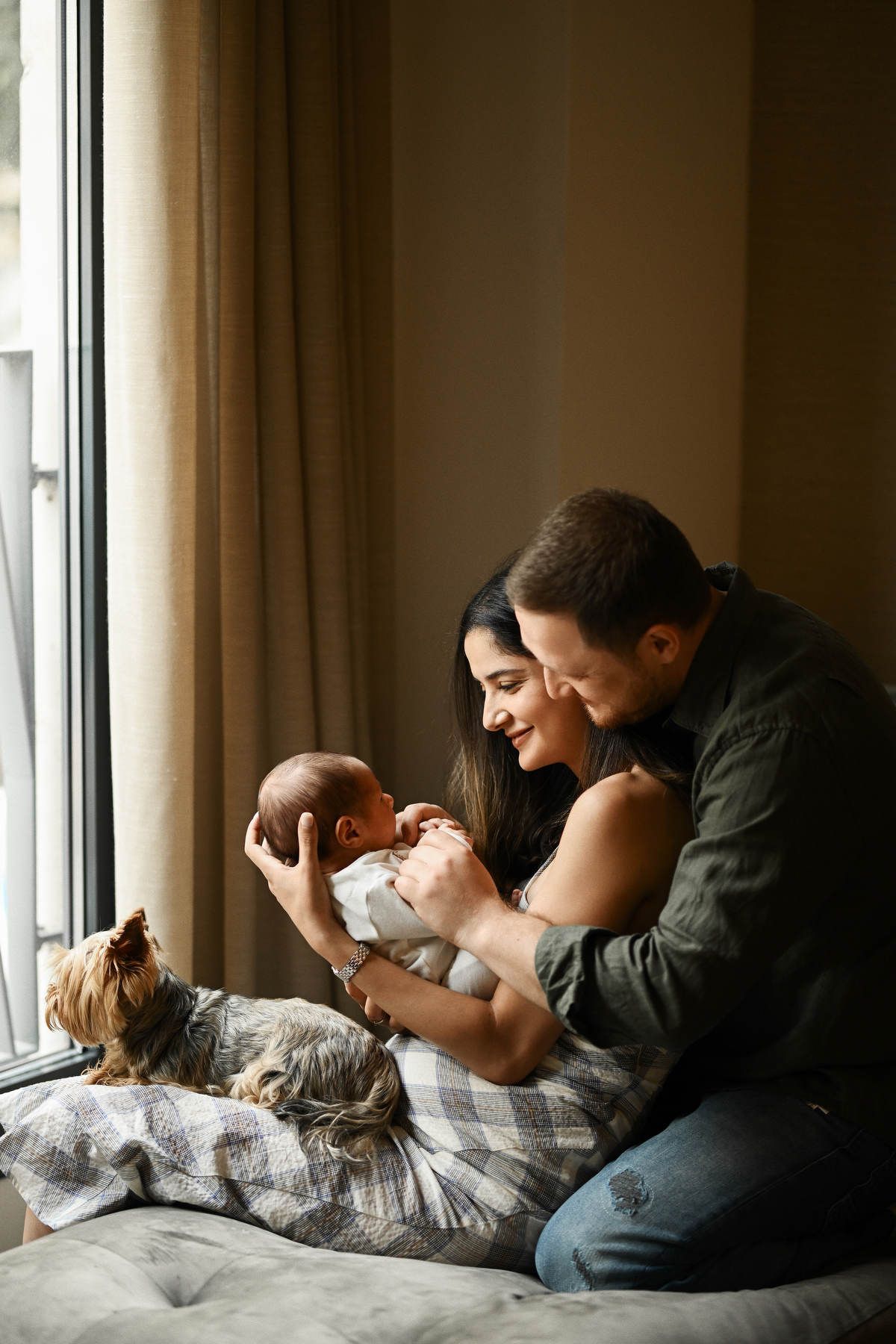 Mother, father, newborn baby, and their dog sitting together by a window during a home photoshoot.