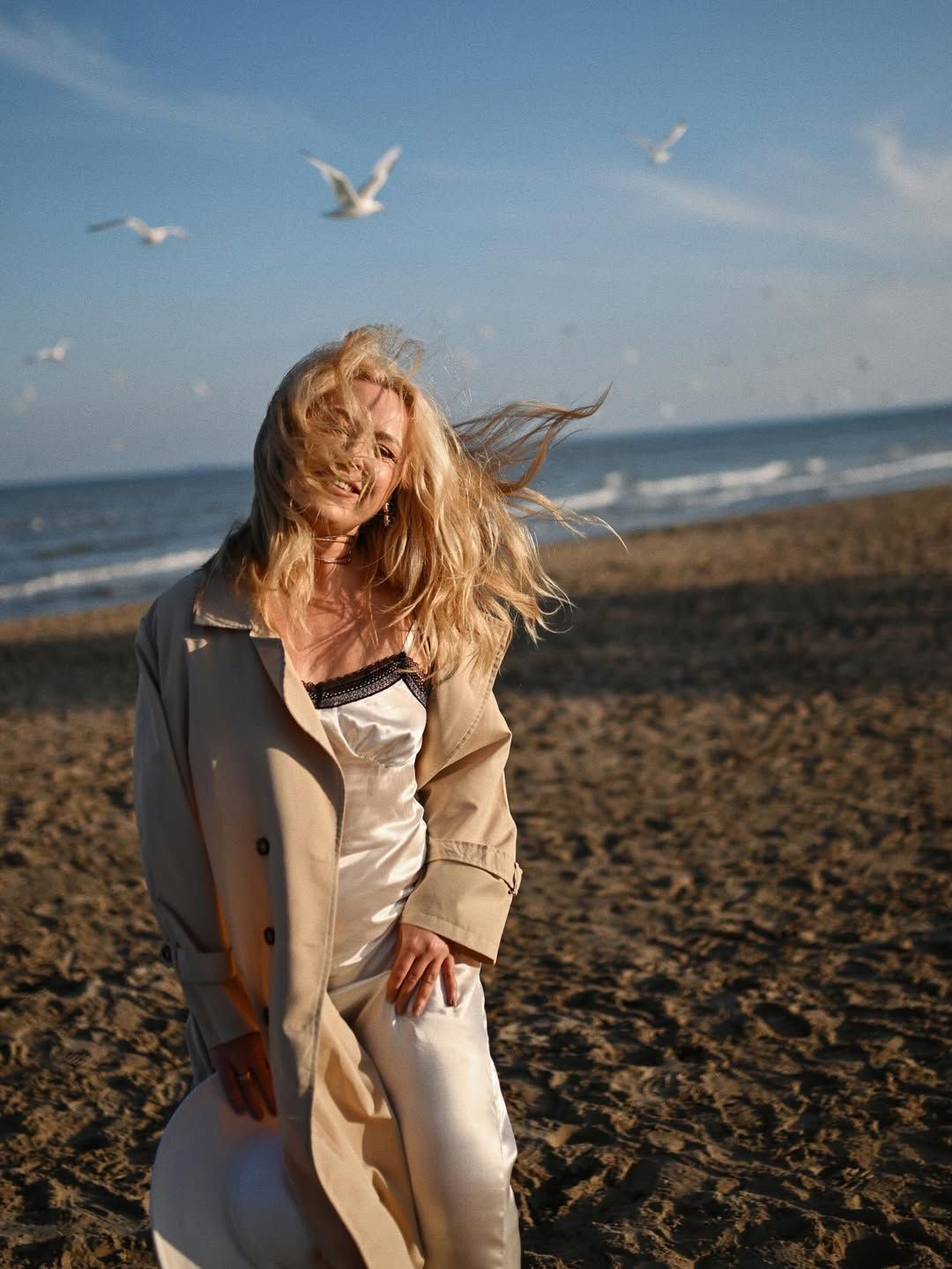 Smiling blonde woman in a silk slip dress and beige trench coat posing on a sandy Costa Blanca beach with seagulls flying behind her.
