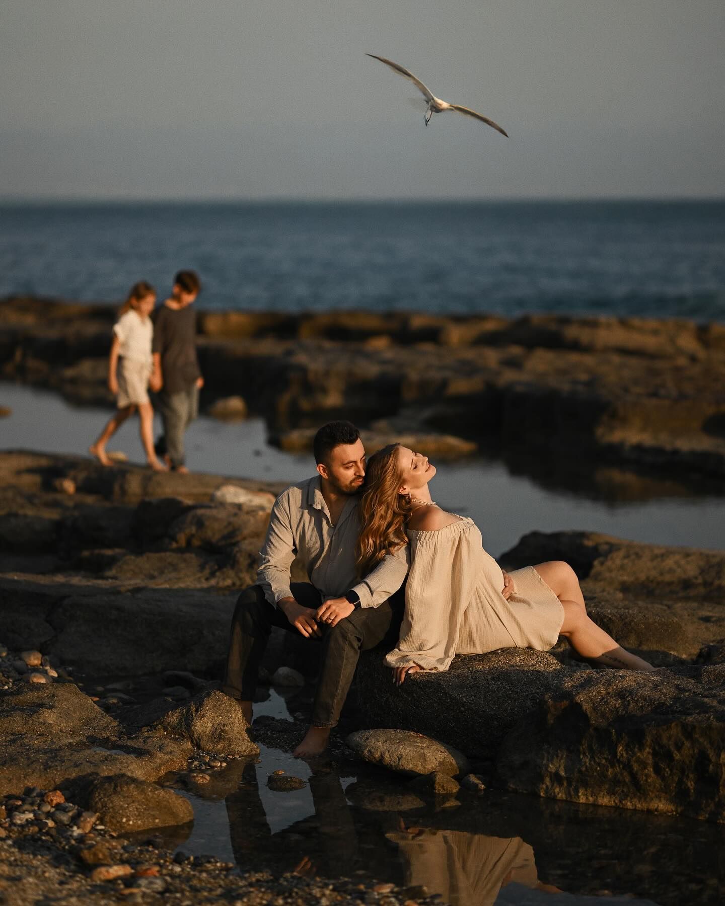 A man and a pregnant woman sitting on coastal rocks at sunset, with children blurred in the background in Costa Blanca.