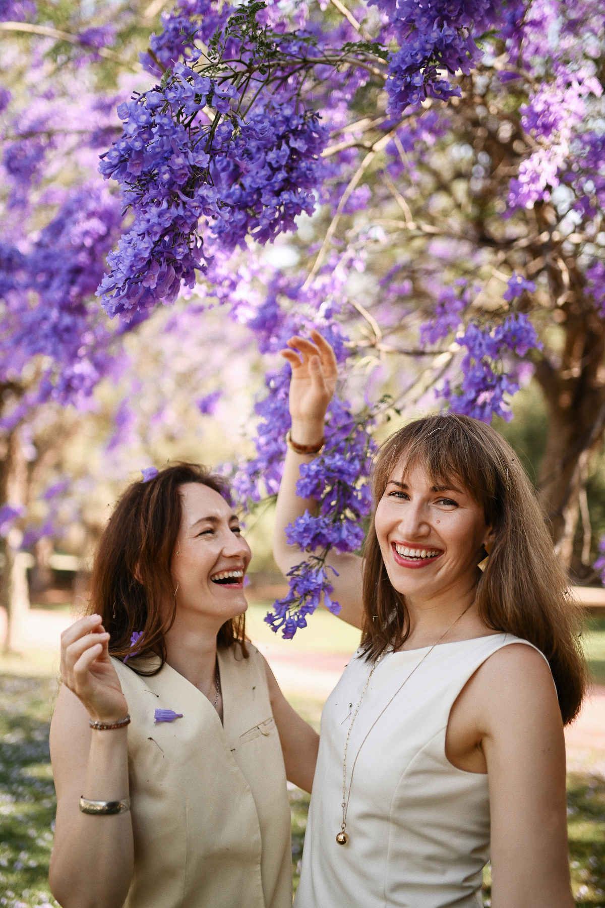 A genuine, candid portrait of two sisters laughing heartily under the bright purple blooms of a Jacaranda tree.