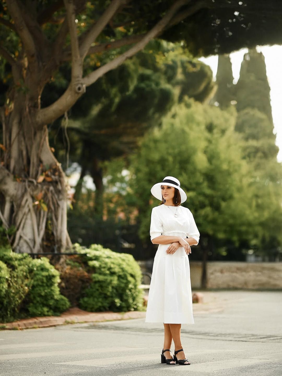 A full-length portrait of a woman in a white dress and matching hat posing gracefully under an old tree in a Valencia park.