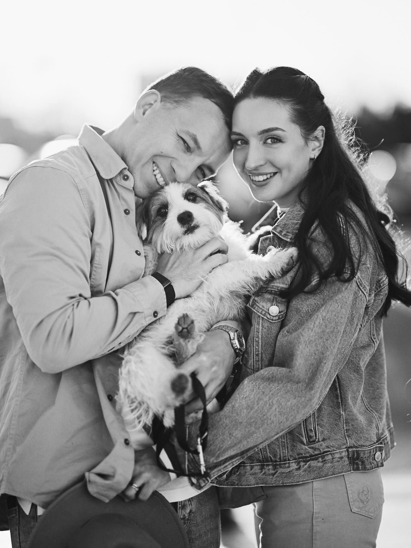 A beautiful black and white close-up portrait of a happy couple smiling and cuddling their cute little dog during an outdoor photoshoot.