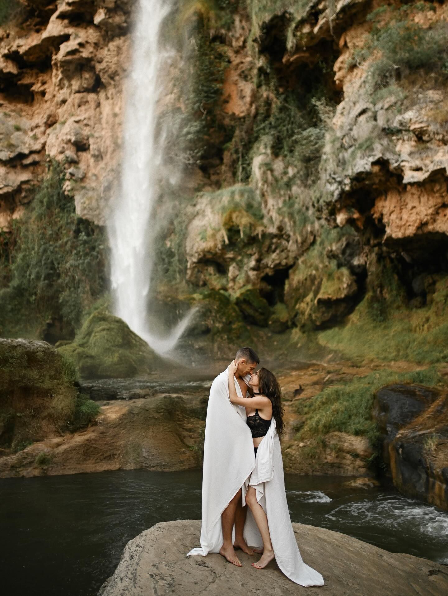 A loving couple wrapped together in a white towel, sharing a tender look on a rock platform in front of a majestic waterfall near Valencia