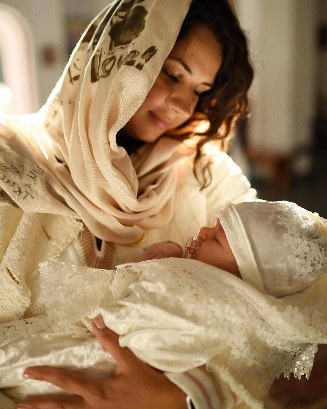 Mother smiling warmly at her sleeping baby wrapped in a white lace blanket after a baptism in Valencia
