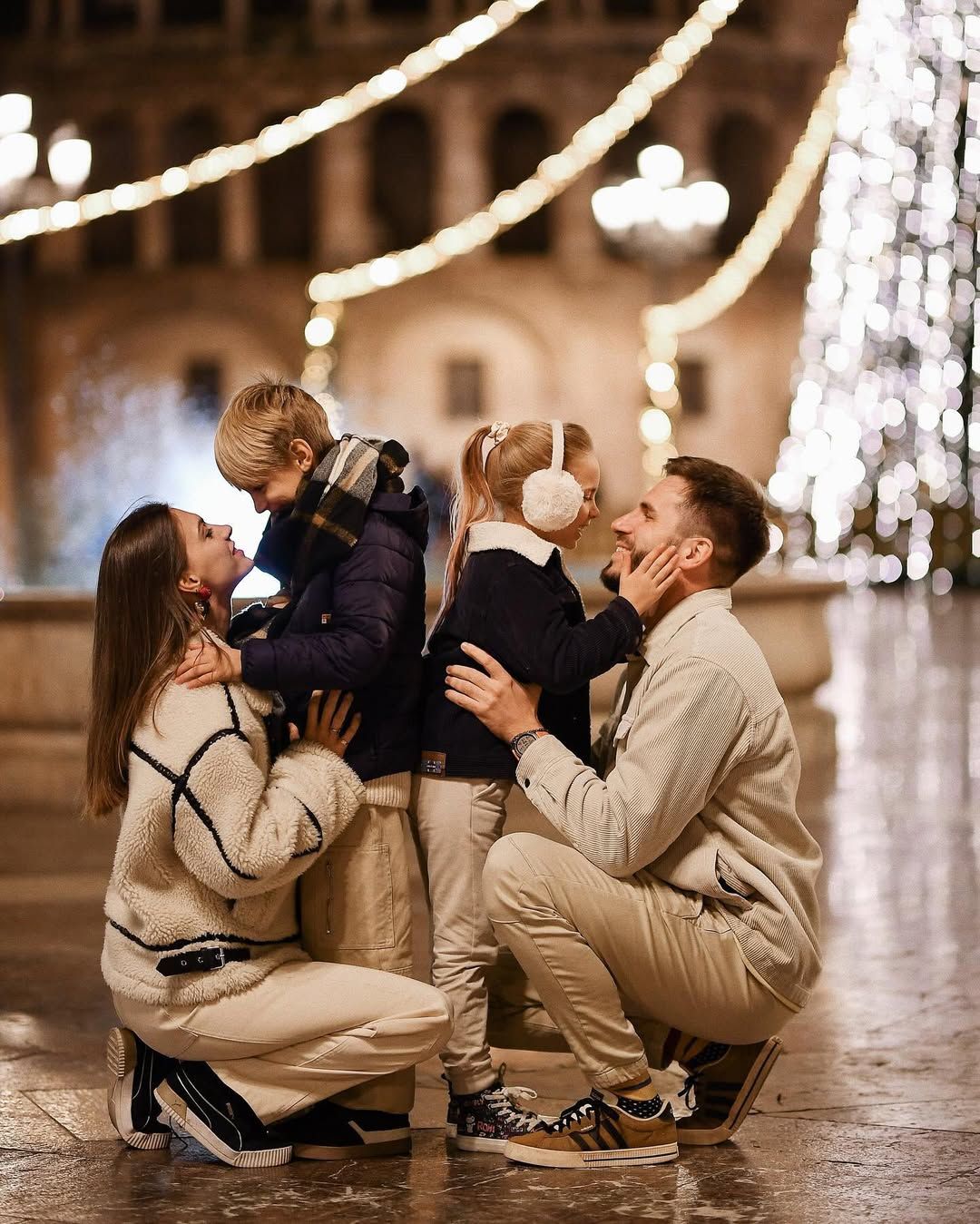 Parents kneeling to hug their smiling kids in a beautifully illuminated Valencia plaza with a Christmas tree behind them