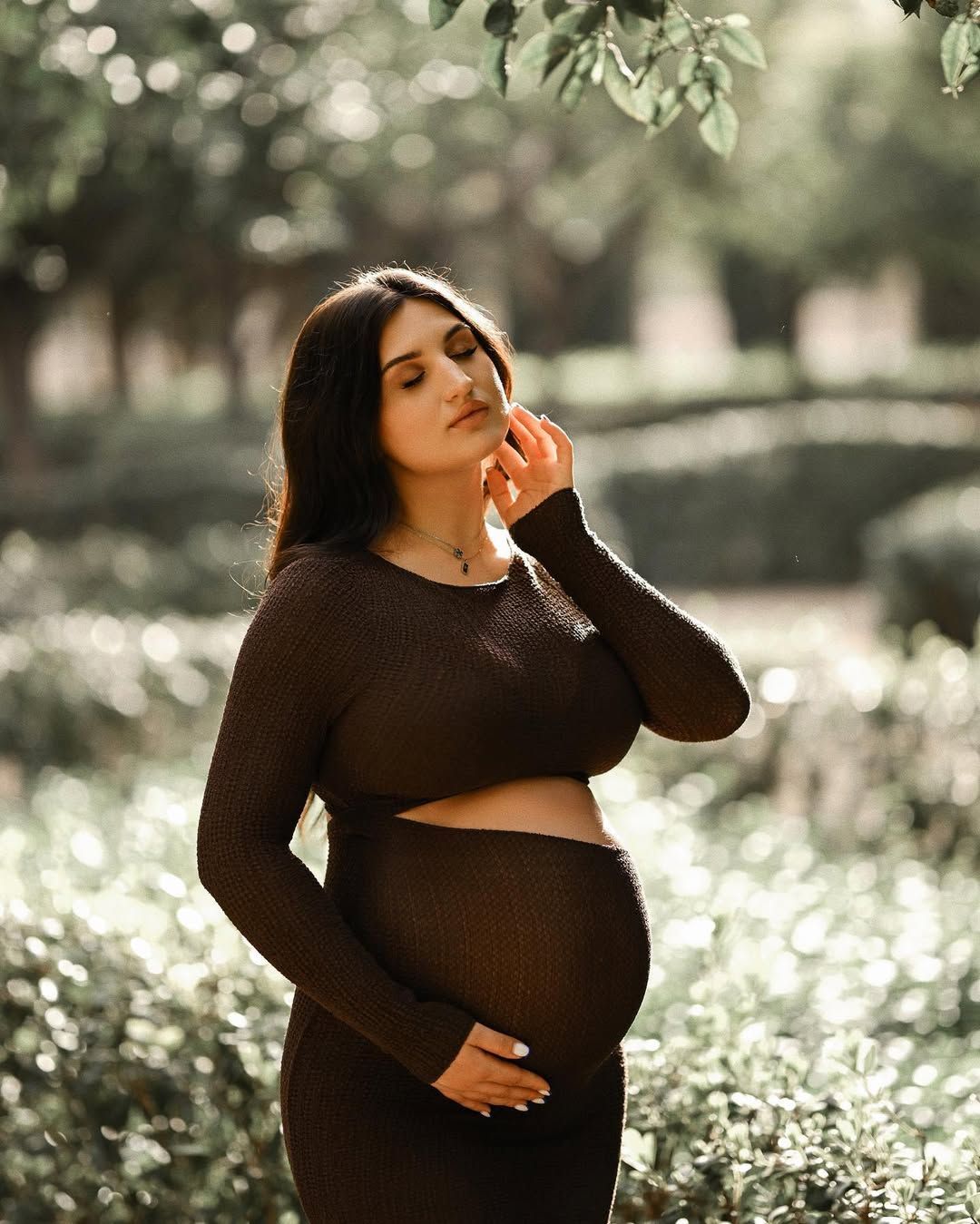 Expecting mother standing with eyes closed in soft dappled light among green foliage at a tranquil Valencia maternity photoshoot