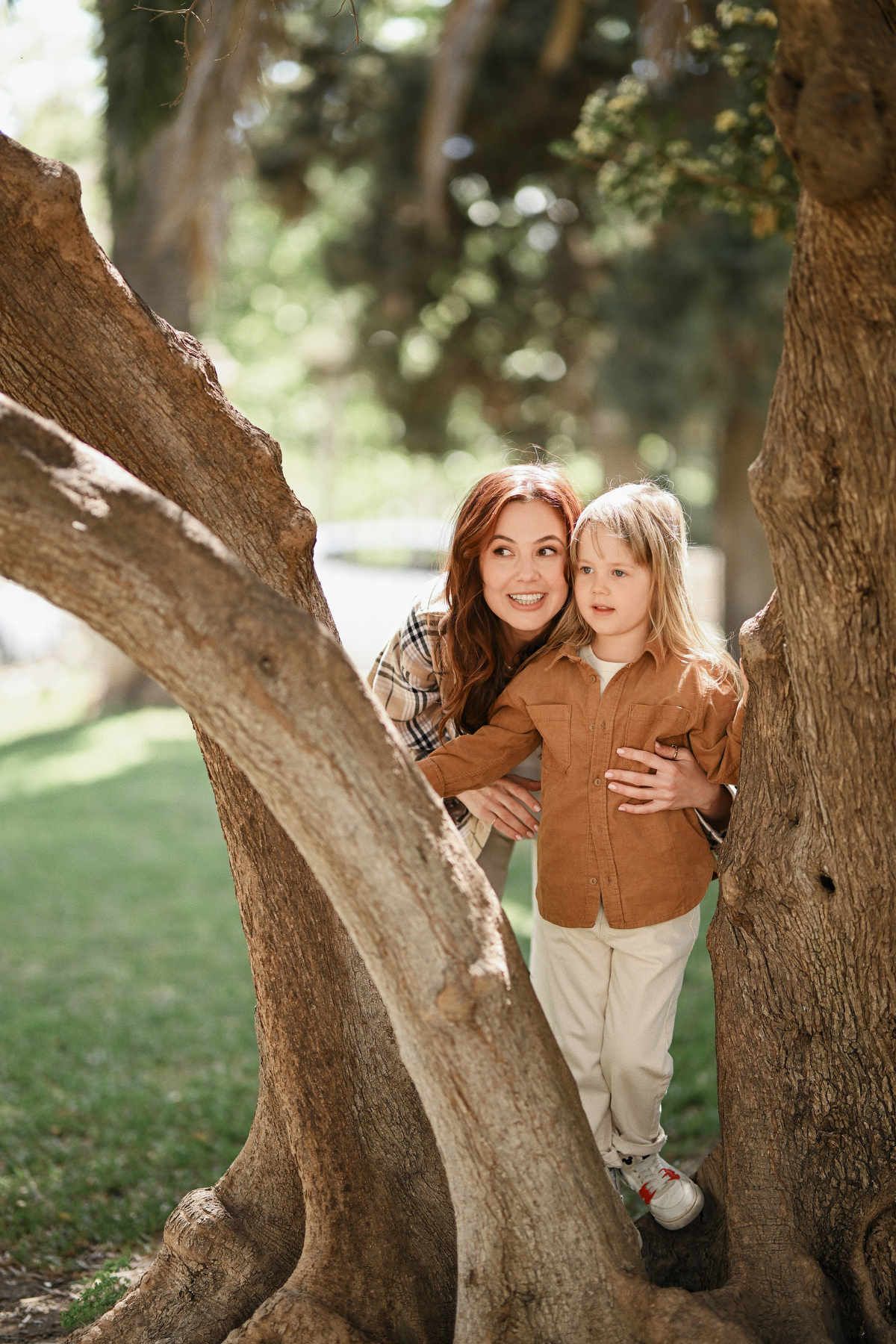 Mother and her young daughter smiling playfully while peeking through thick wooden trunks in Viveros Park.