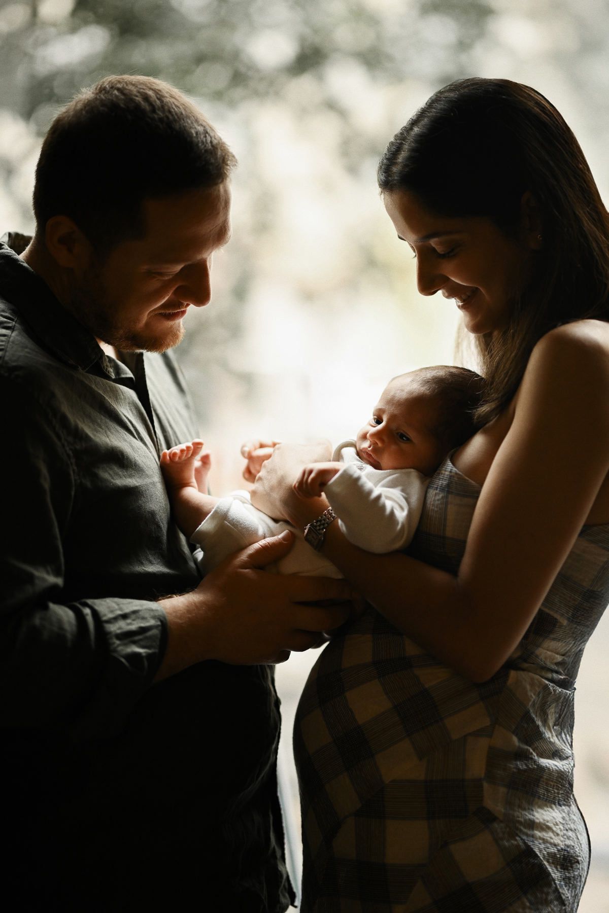 Backlit lifestyle portrait of a mother and father holding their newborn baby together by the window.