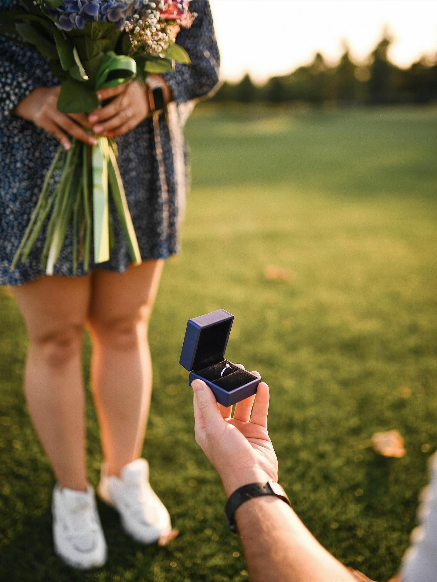 Man holding a blue velvet ring box, proposing to his girlfriend in a sunlit park during a professional proposal session in Valencia.