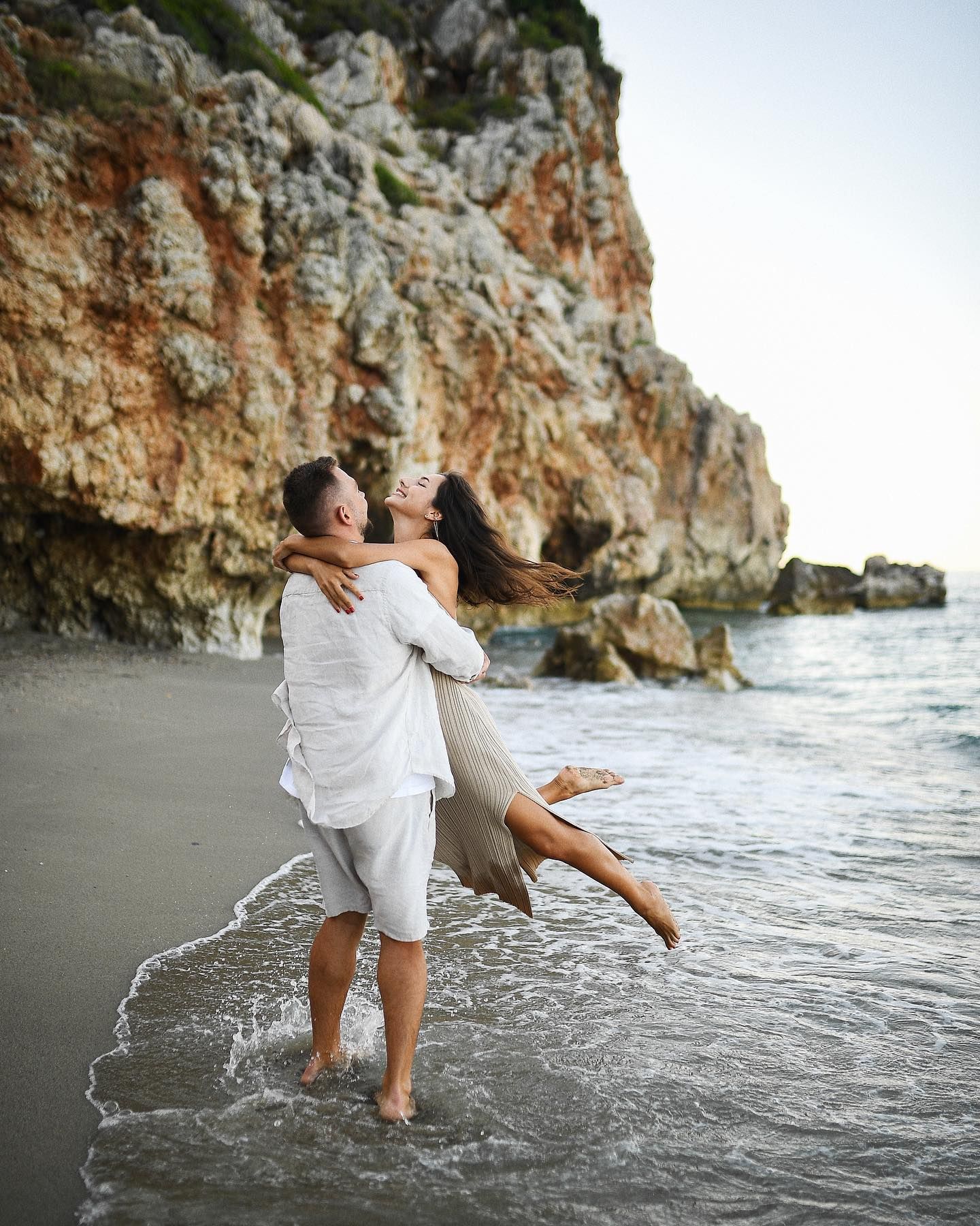 A man playfully lifts his wife on the beach during their romantic honeymoon photoshoot in Valencia, with a stunning coastline background.