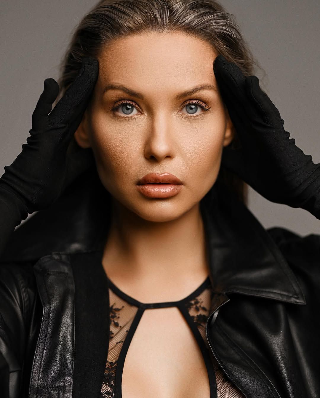 A striking close-up studio portrait of a blonde woman holding her head with black gloves, showcasing clean fashion makeup.