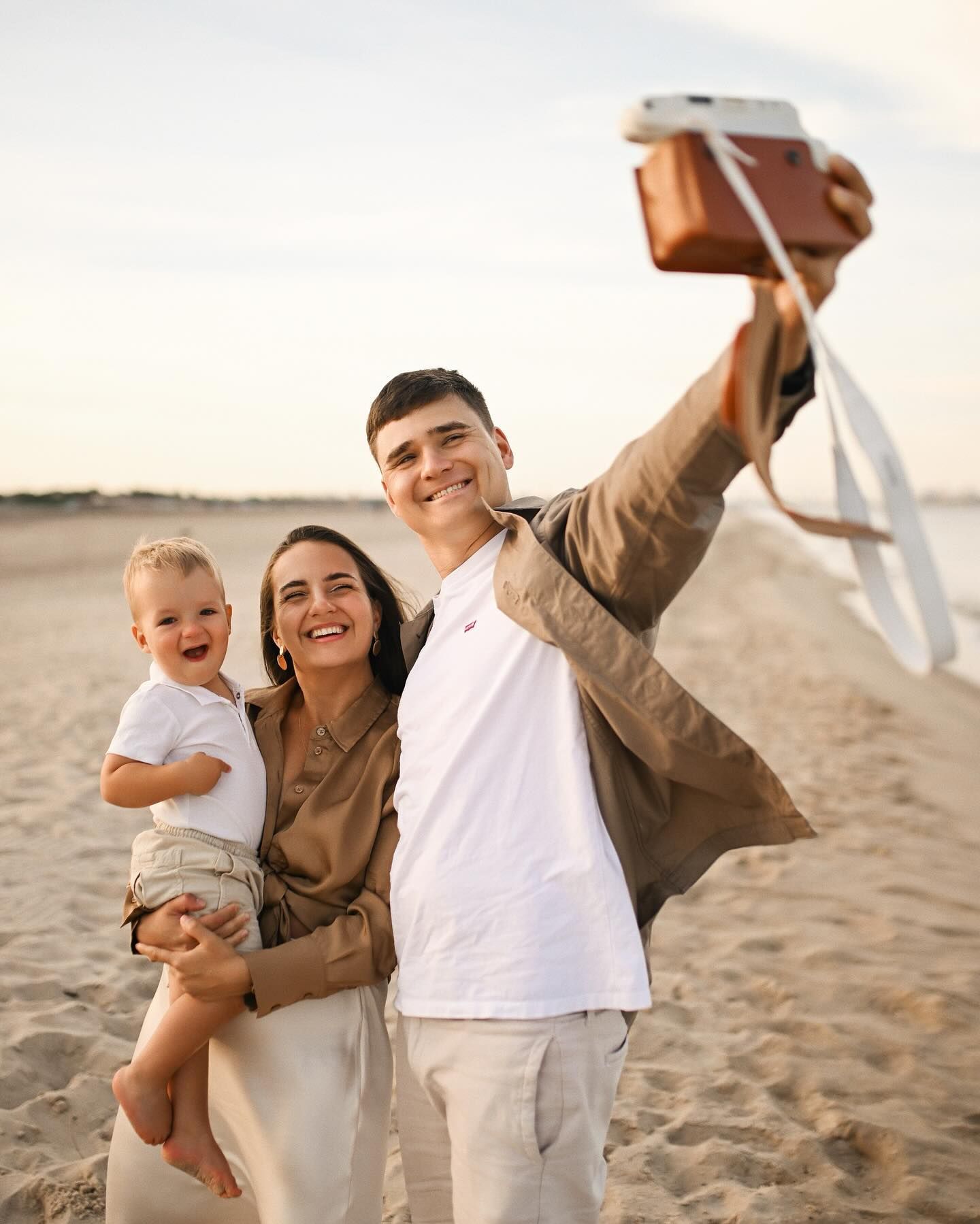Happy family holding a vintage camera while taking a selfie on a sandy beach during a relaxed sunset photoshoot in Valencia.