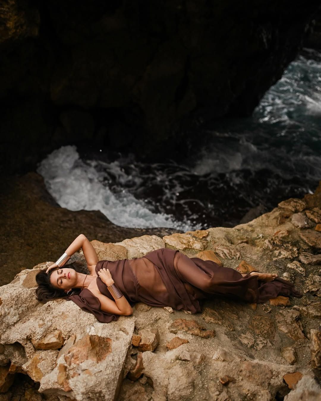 Cinematic portrait of a woman in a sheer dress reclining on rugged rocks at Cala Moraig beach.