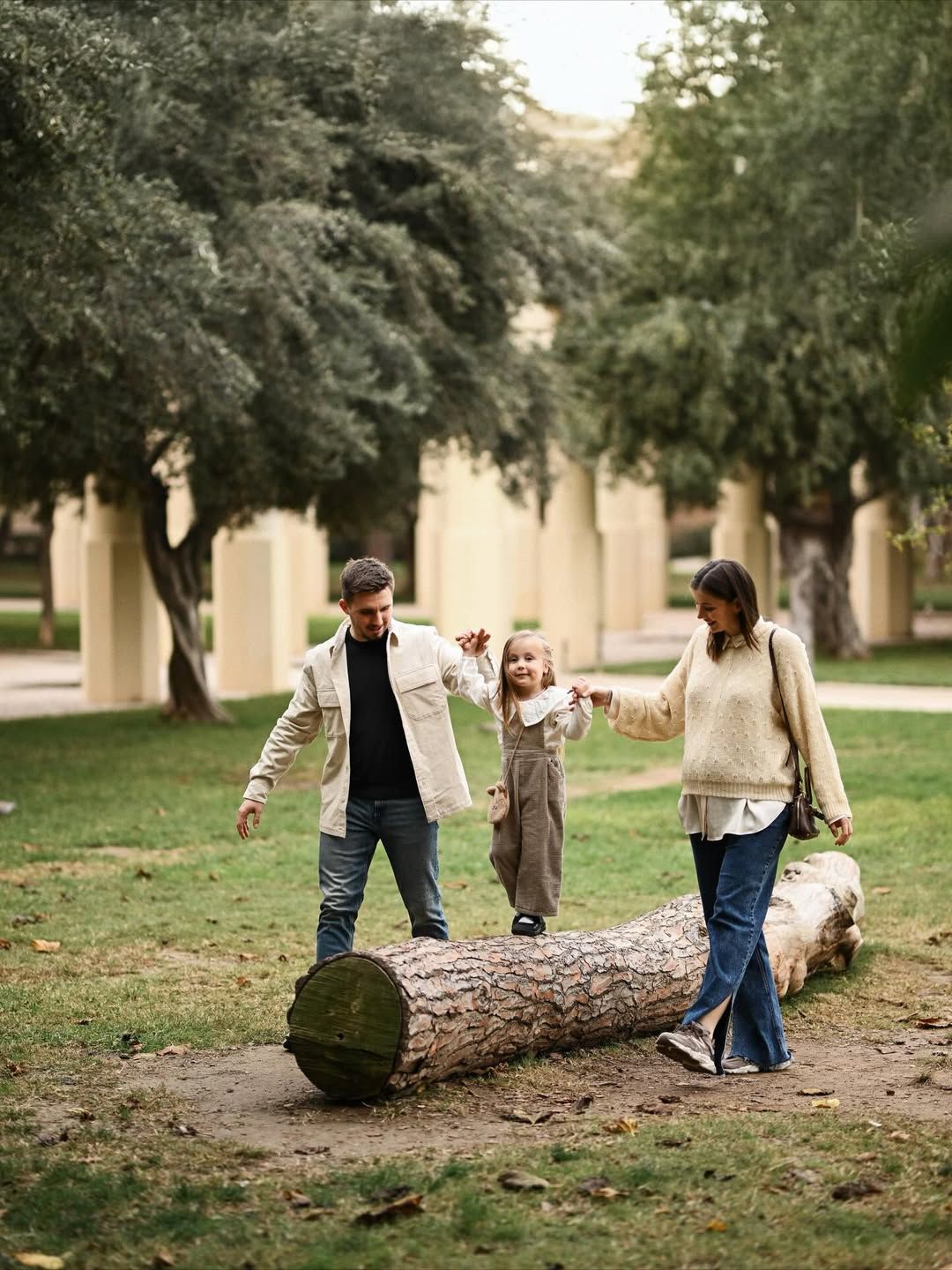 Family maternity session in Turia Park, Valencia with parents helping their daughter balance on a fallen tree surrounded by greenery.