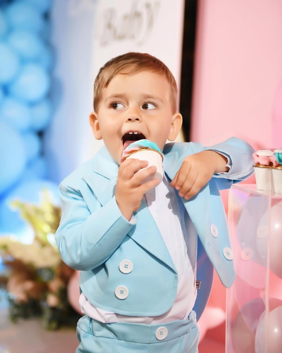 Cute little boy in a blue suit happily eating a cupcake from the styled candy bar at a gender reveal party in Valencia.