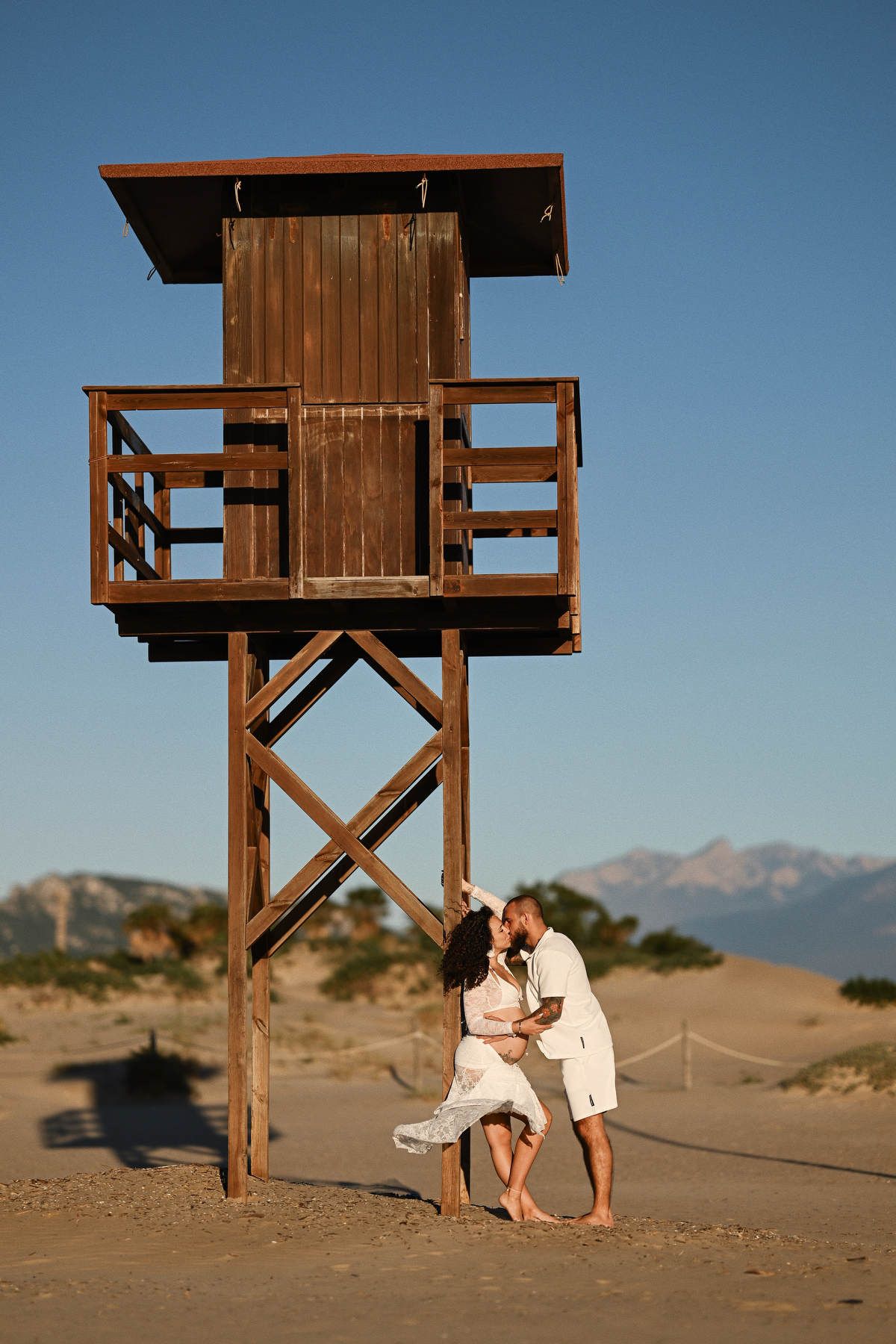 A pregnant woman and her partner sharing a kiss by a wooden lifeguard tower on a beautiful sandy beach.
