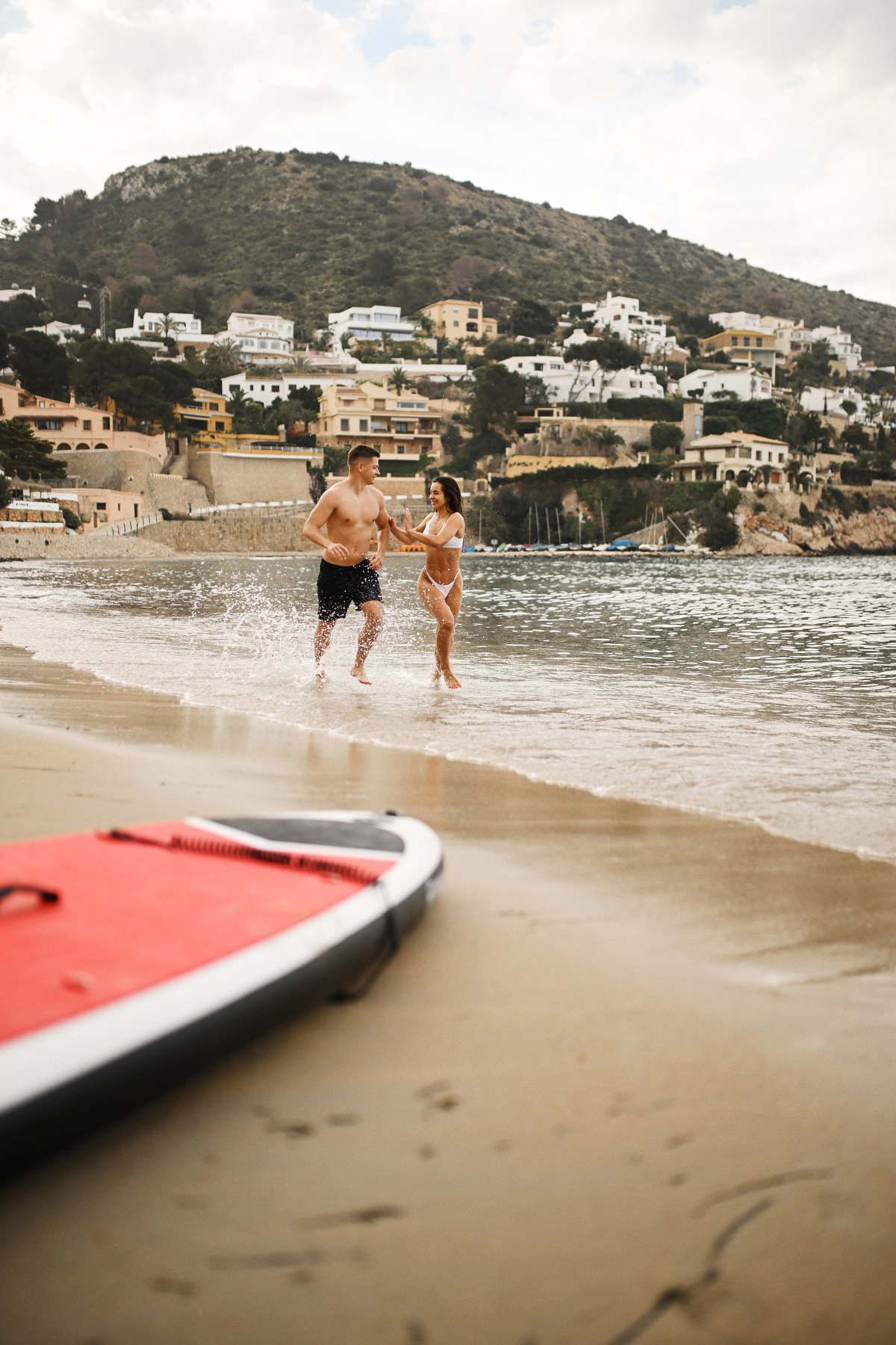 A joyful couple splashing out of the sea at Playa del Portet, with Moraira's iconic hillside villas behind them