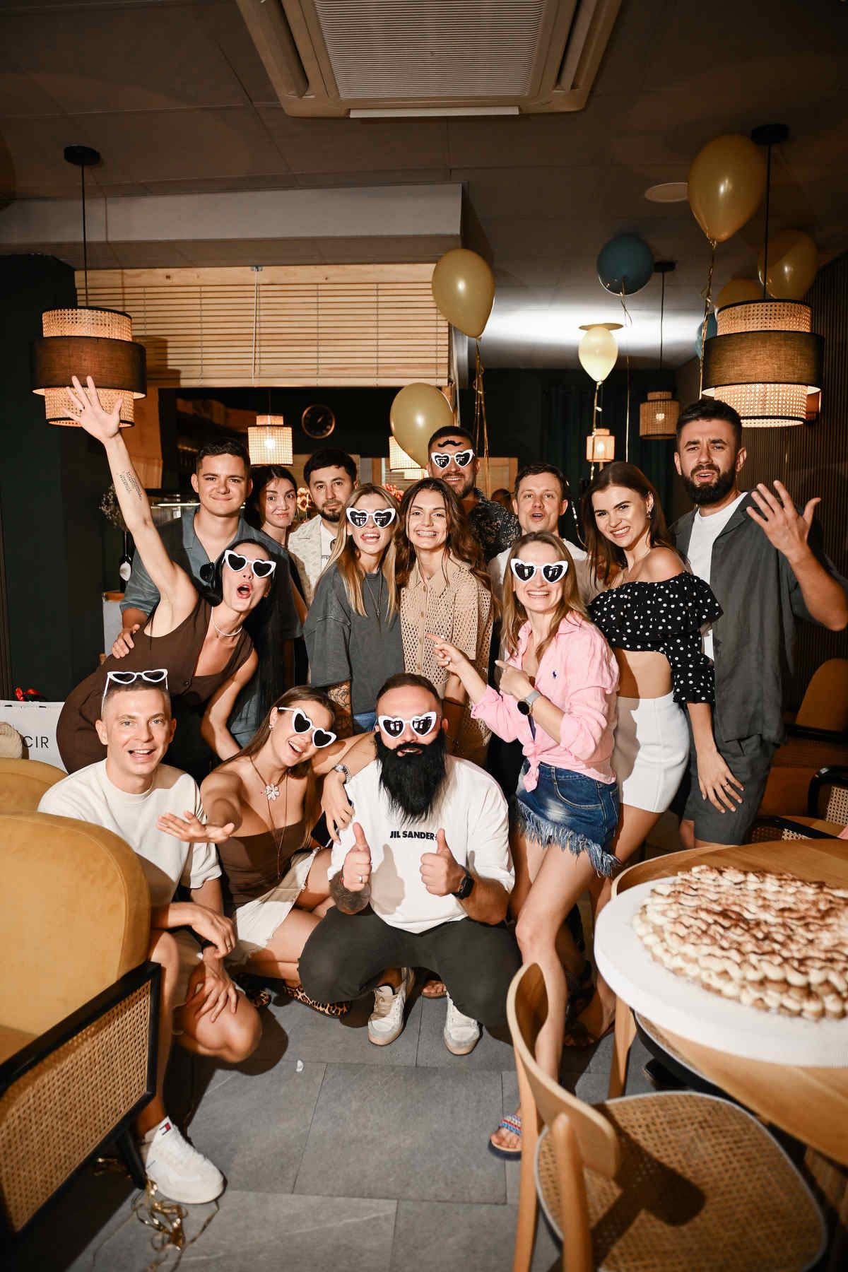 Large group of friends in heart-shaped glasses posing for a fun group photo at a birthday party with balloons.