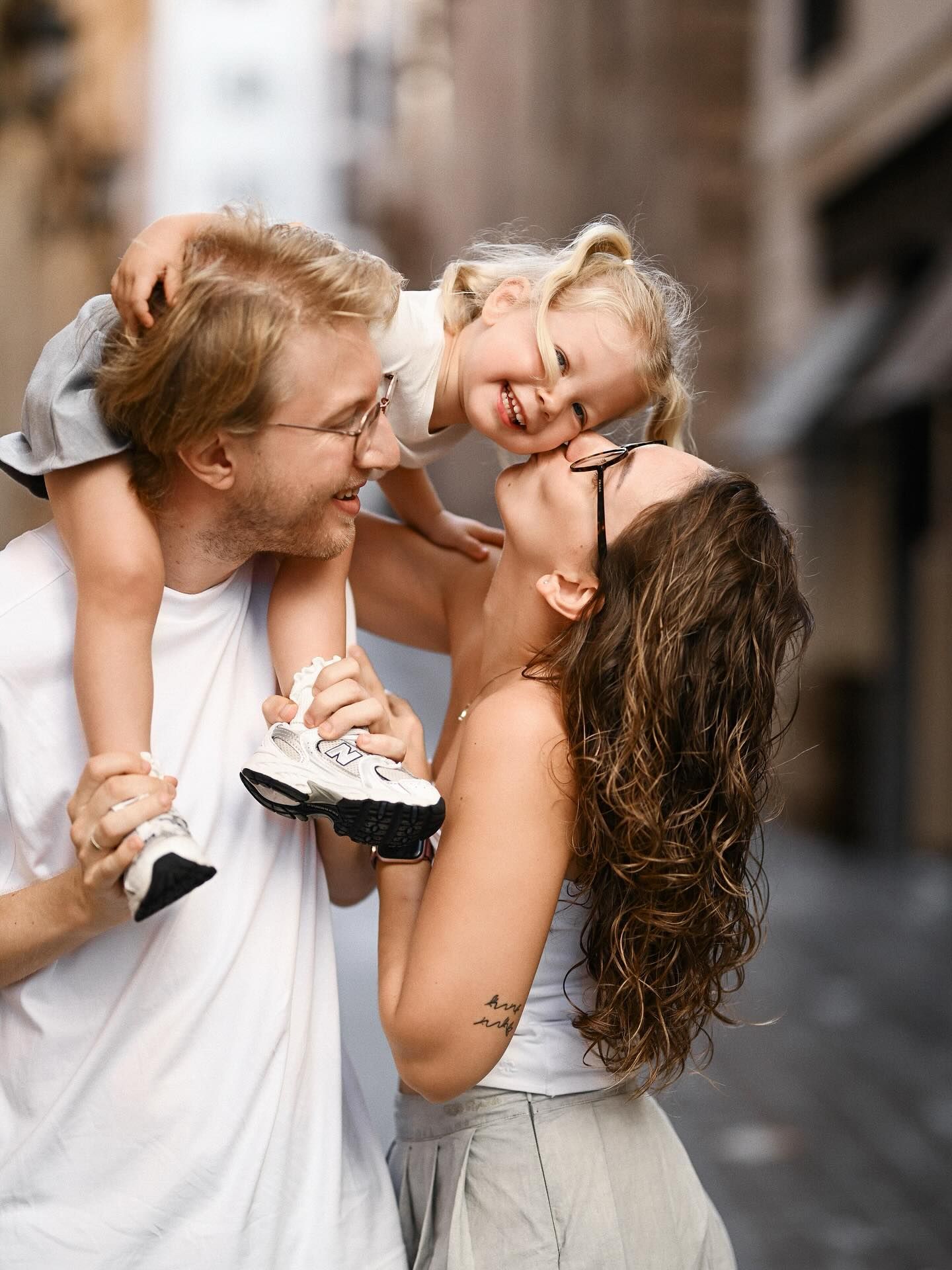 A young family walking down a picturesque city street as the father carries their smiling little daughter on his shoulders.