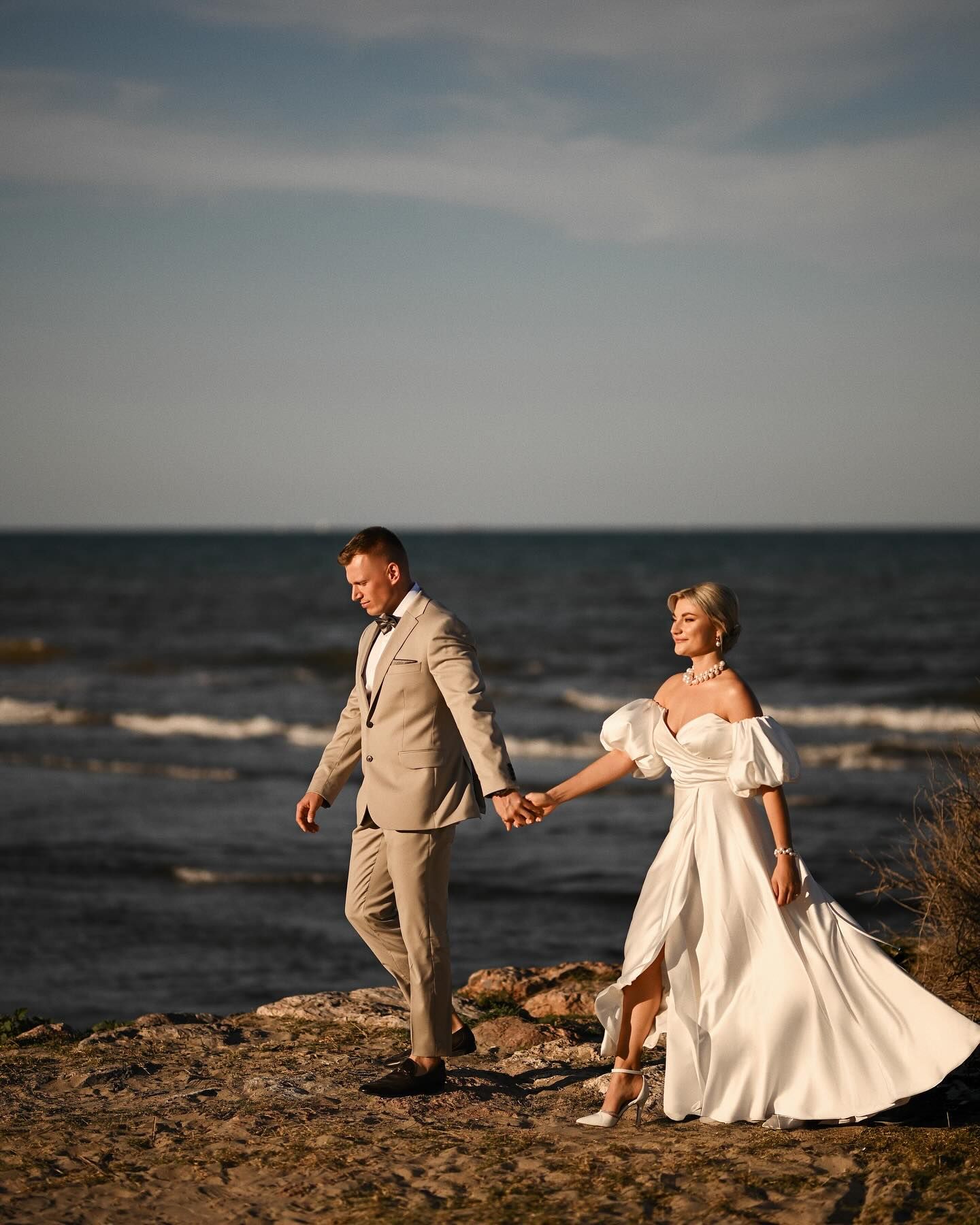 An elegant couple in wedding outfits walks along the cinematic coastline during their romantic Valencia elopement session at golden hour.