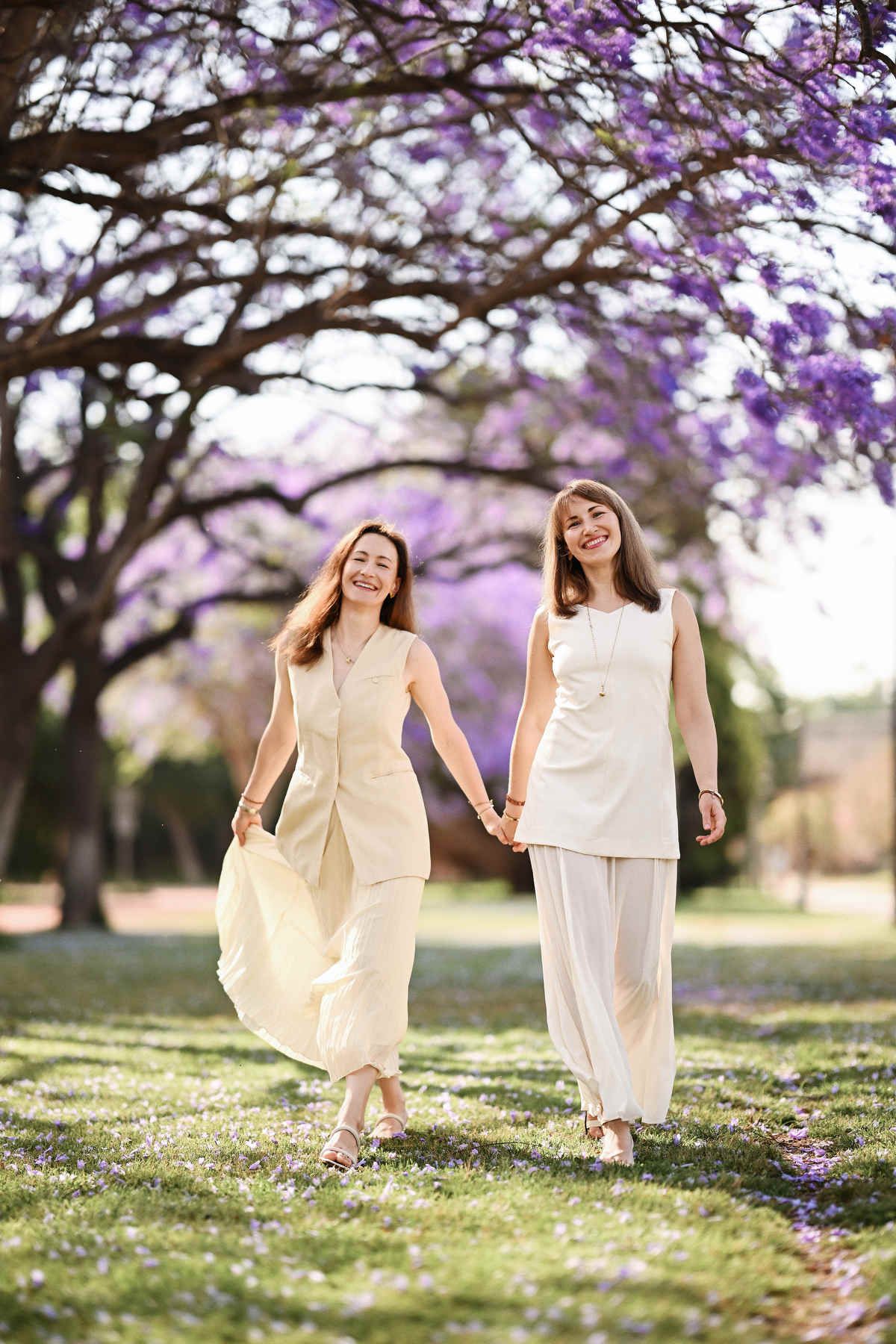 Full-length lifestyle portrait of two happy sisters walking hand in hand through a park scattered with fallen petals.