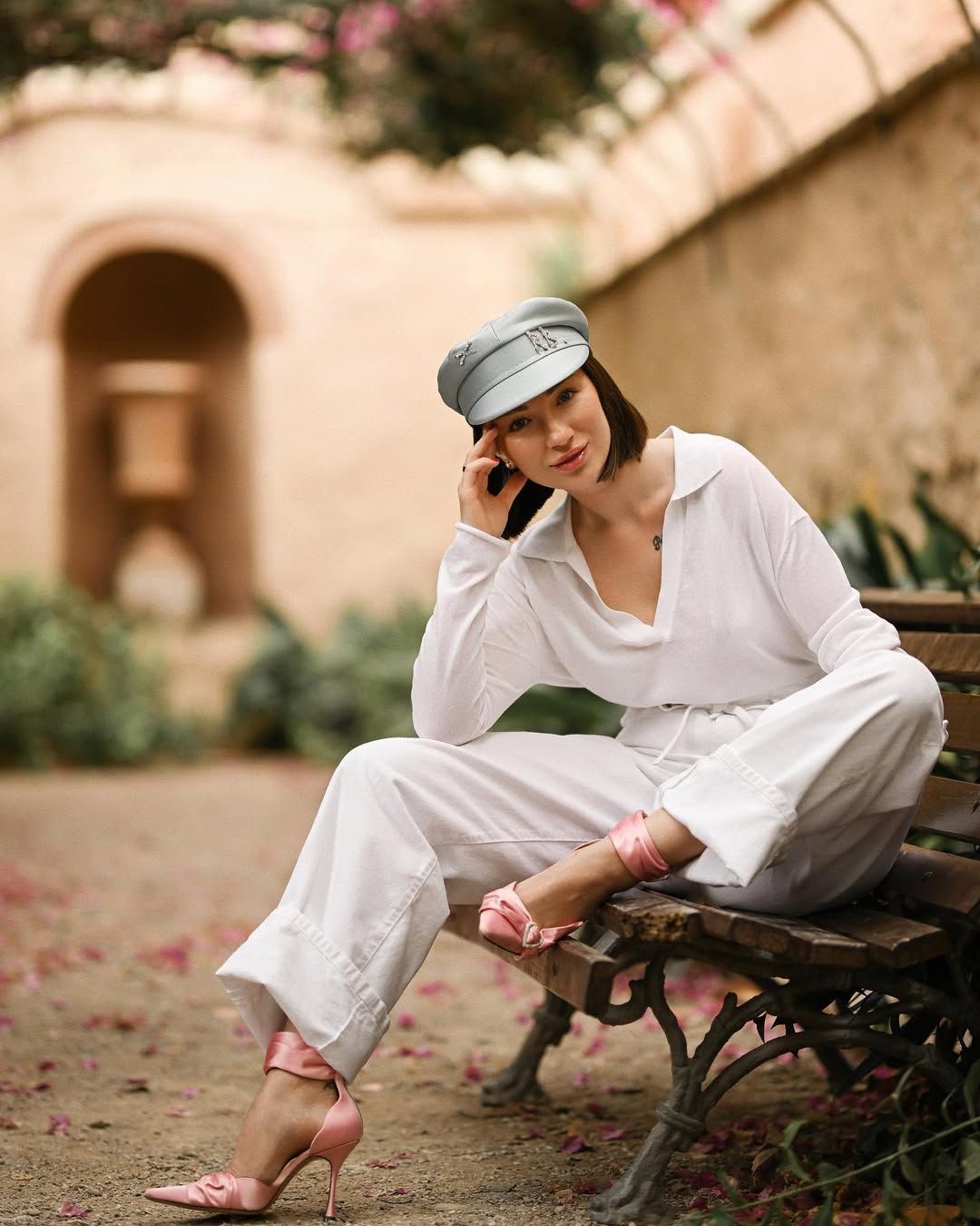 A woman in a white suit and leather cap sitting on a wooden bench in Monforte Gardens, Valencia.