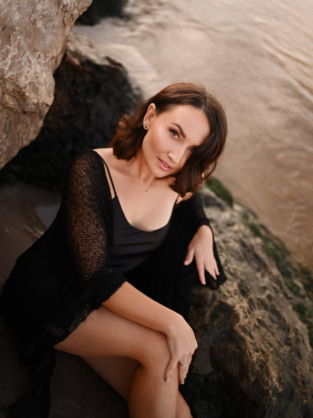 High-angle women's portrait on a Valencia coast, woman in black sitting relaxed against a large rock near the water edge.
