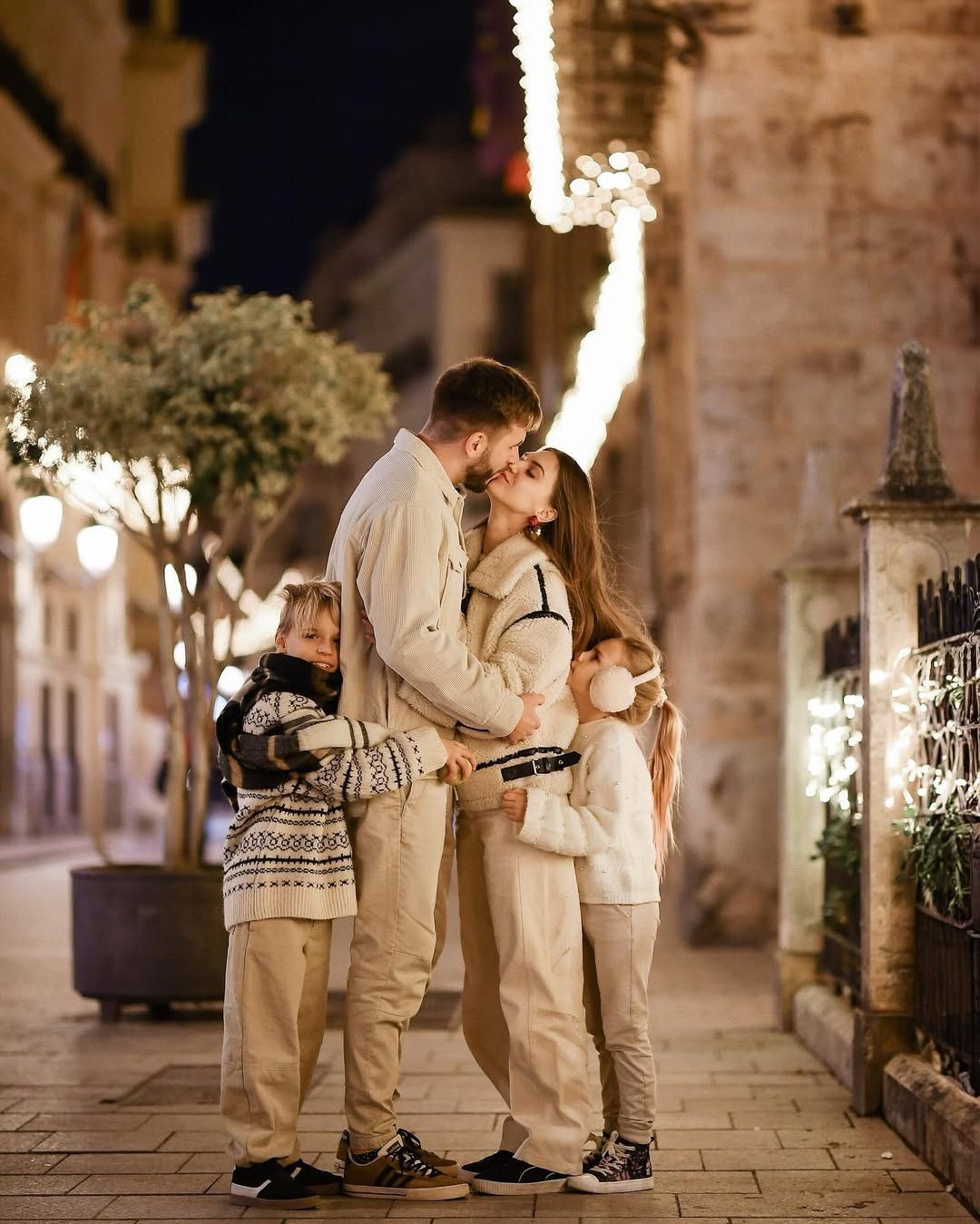 Parents kissing while their children lovingly hug them on a festive, light-filled street during a Valencia holiday session