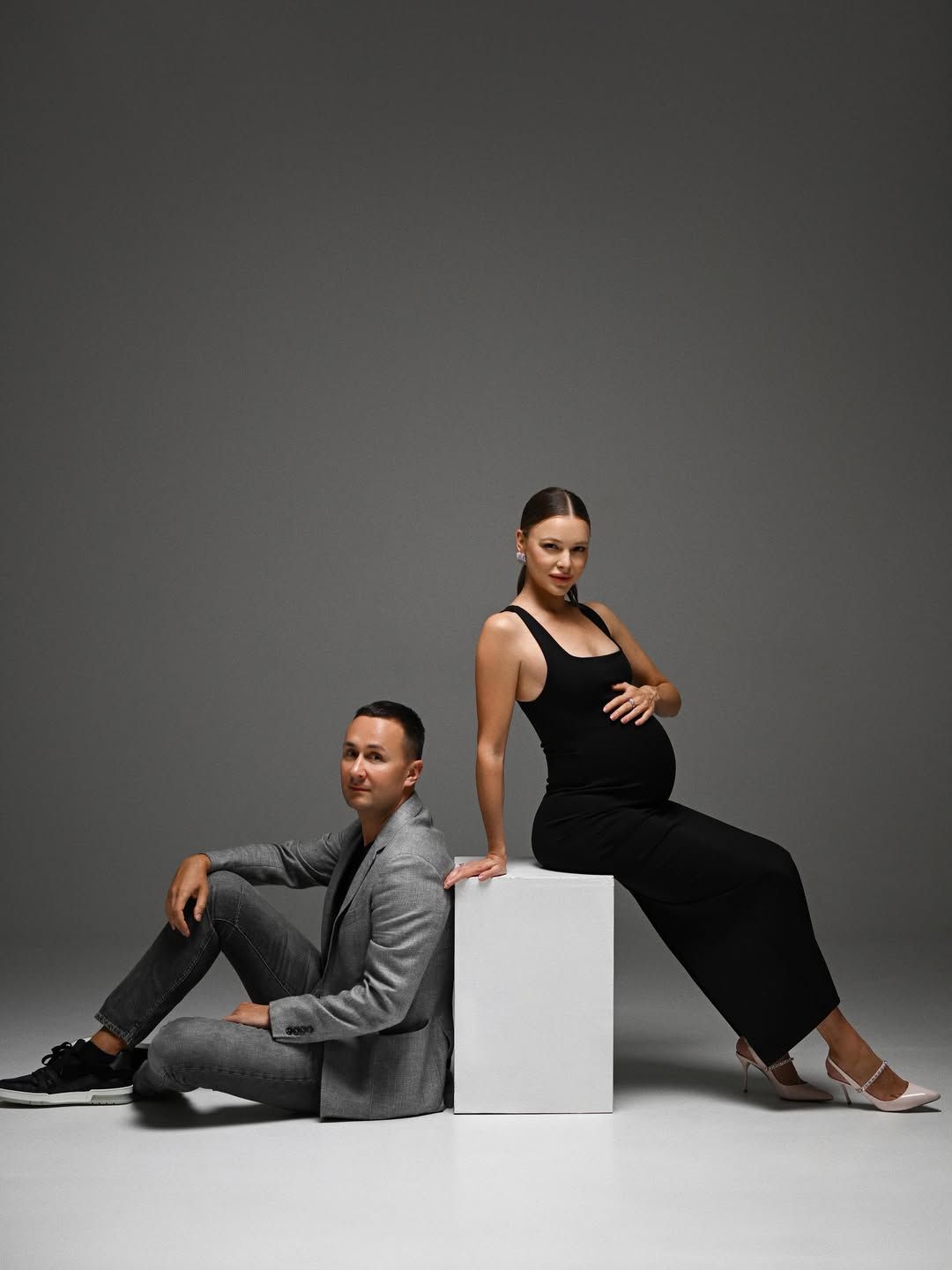A stylish pregnant woman sitting on a white studio cube with her partner posing on the floor against a neutral grey background