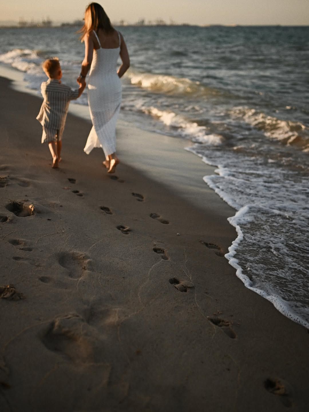 A mom and her boy walking hand in hand towards the sea, leaving footprints in the wet sand at El Saler beach.