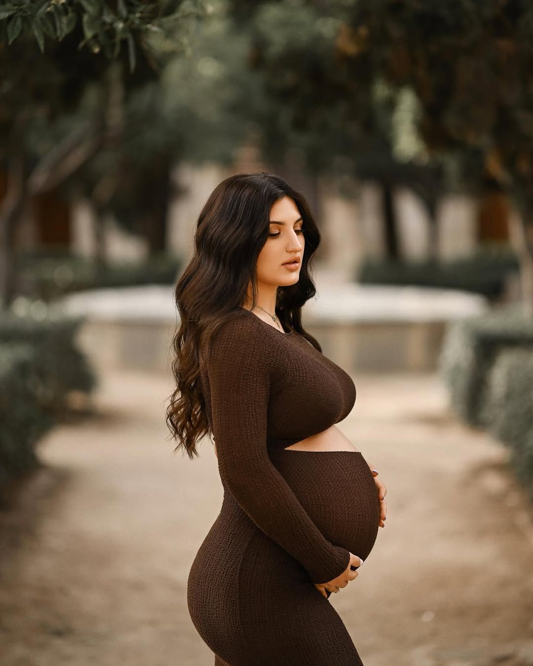 Expecting mother walking along a quiet tree-lined path in Jardines del Real, cradling her bump during a Valencia pregnancy session