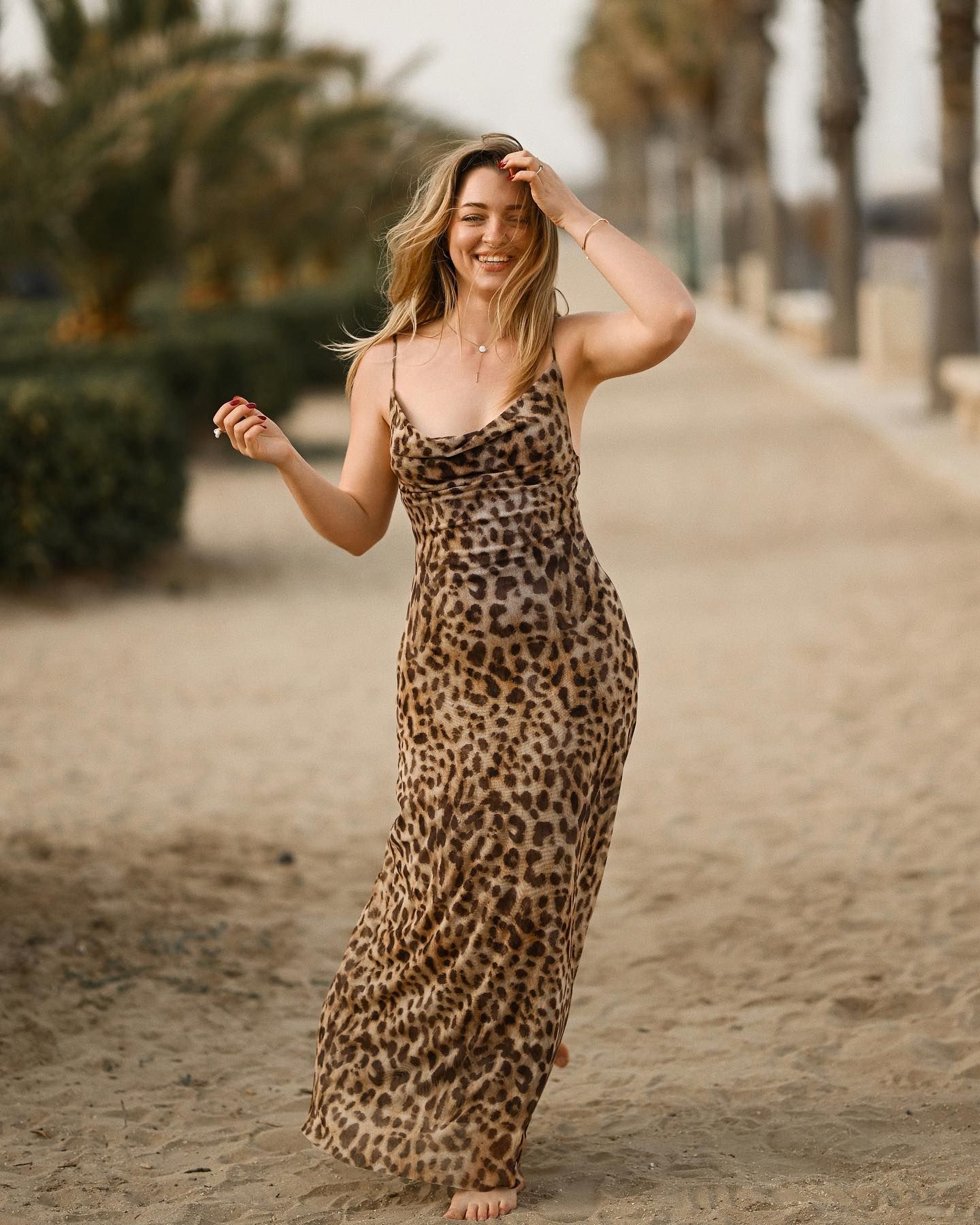 A happy blonde woman in a leopard print dress running barefoot on the sand near palm trees in Alicante.