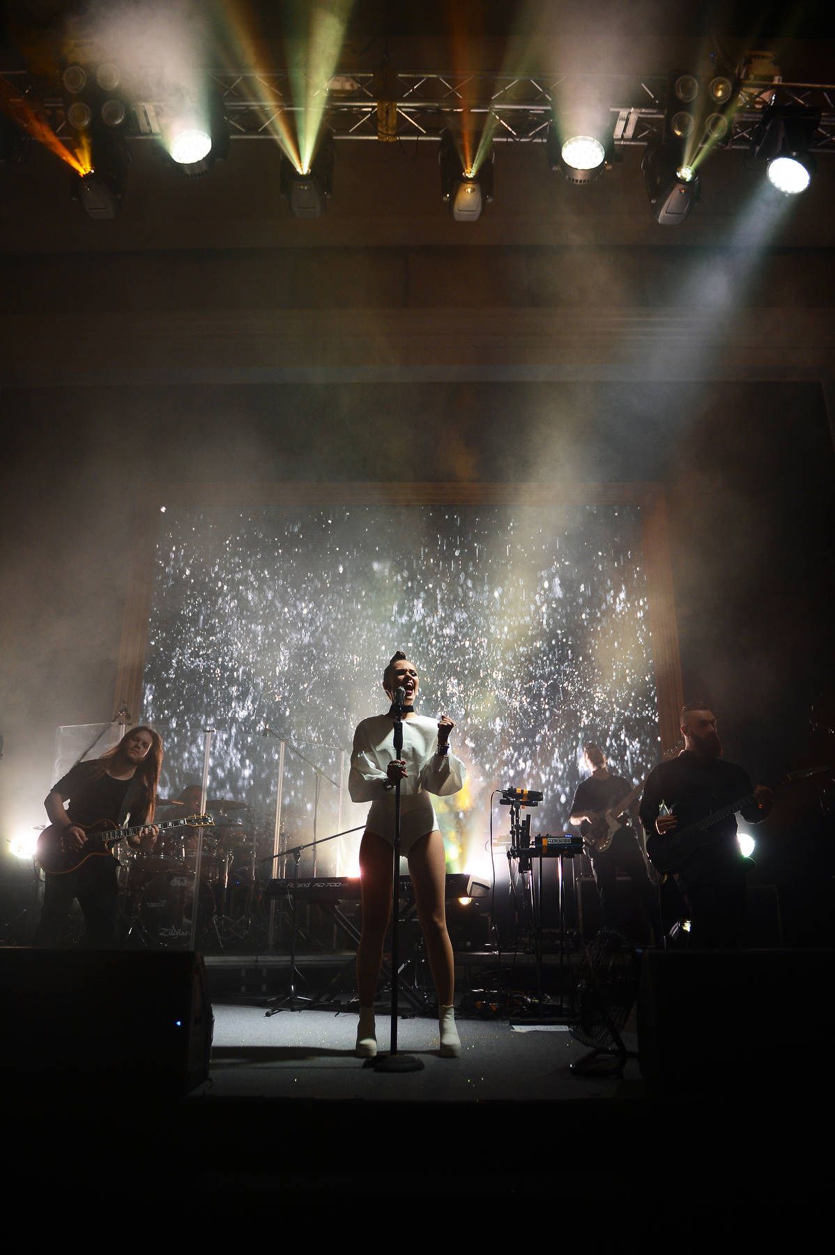 A female singer in a white outfit performing live on stage with her band under dynamic spotlights and a screen backdrop.