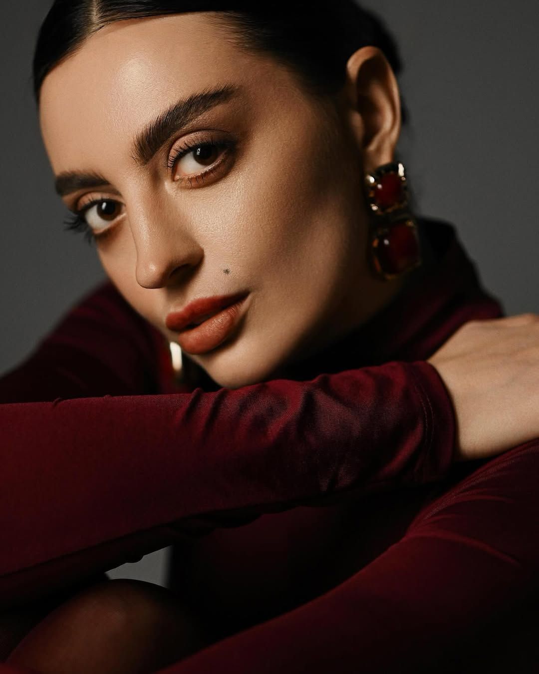 A dramatic close-up studio portrait of a woman in a dark red turtleneck, focusing on her intense gaze and elegant makeup.