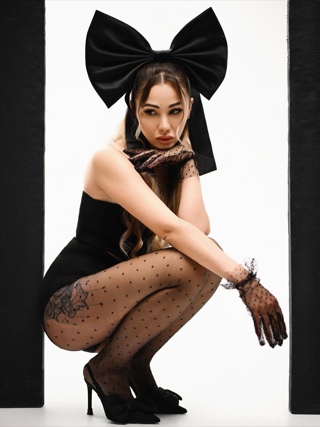 A creative studio portrait showing a woman crouching between black panels, styled with a giant black satin head bow and lace gloves.
