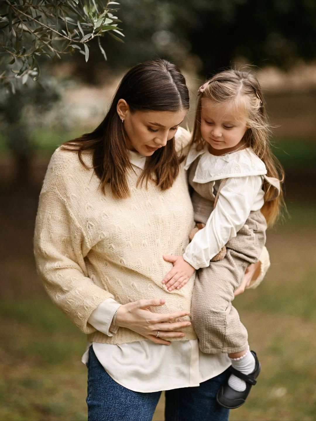 Expecting mother holding her daughter as both gently rest their hands on the baby bump beneath the trees in Turia Park, Valencia.