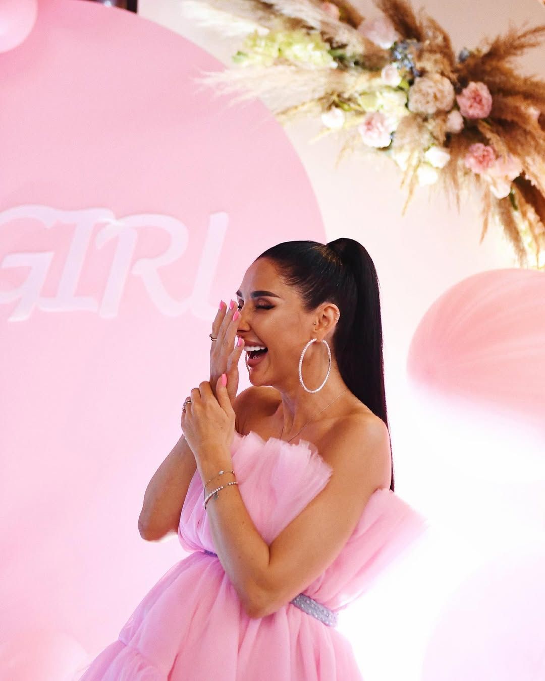 Laughing pregnant woman in a pink dress reacting with joy at her styled indoor gender reveal party and baby shower in Valencia.